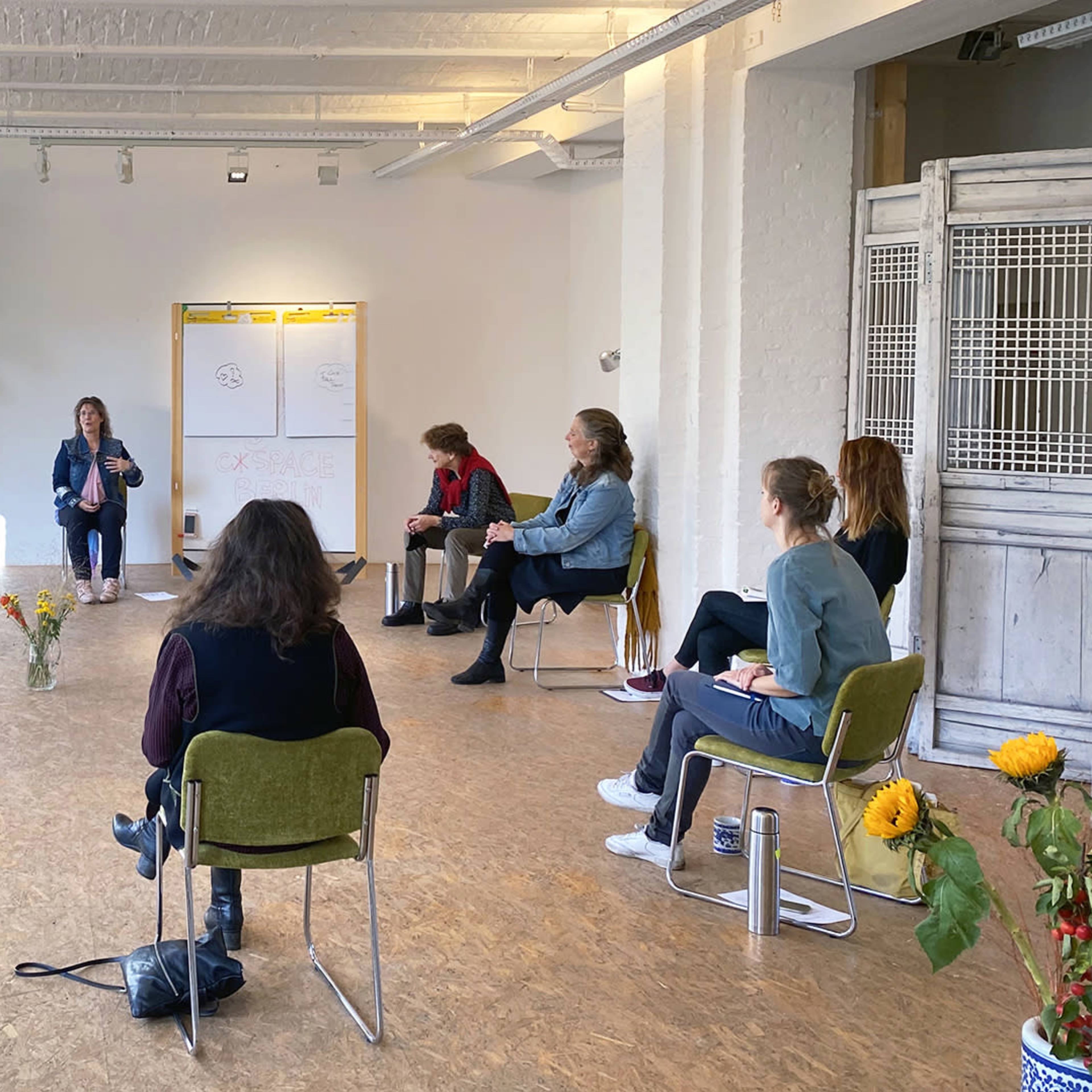 A group of people sit in chairs arranged in a circle as a speaker presents ideas in a well-lit room with large windows and plants.
