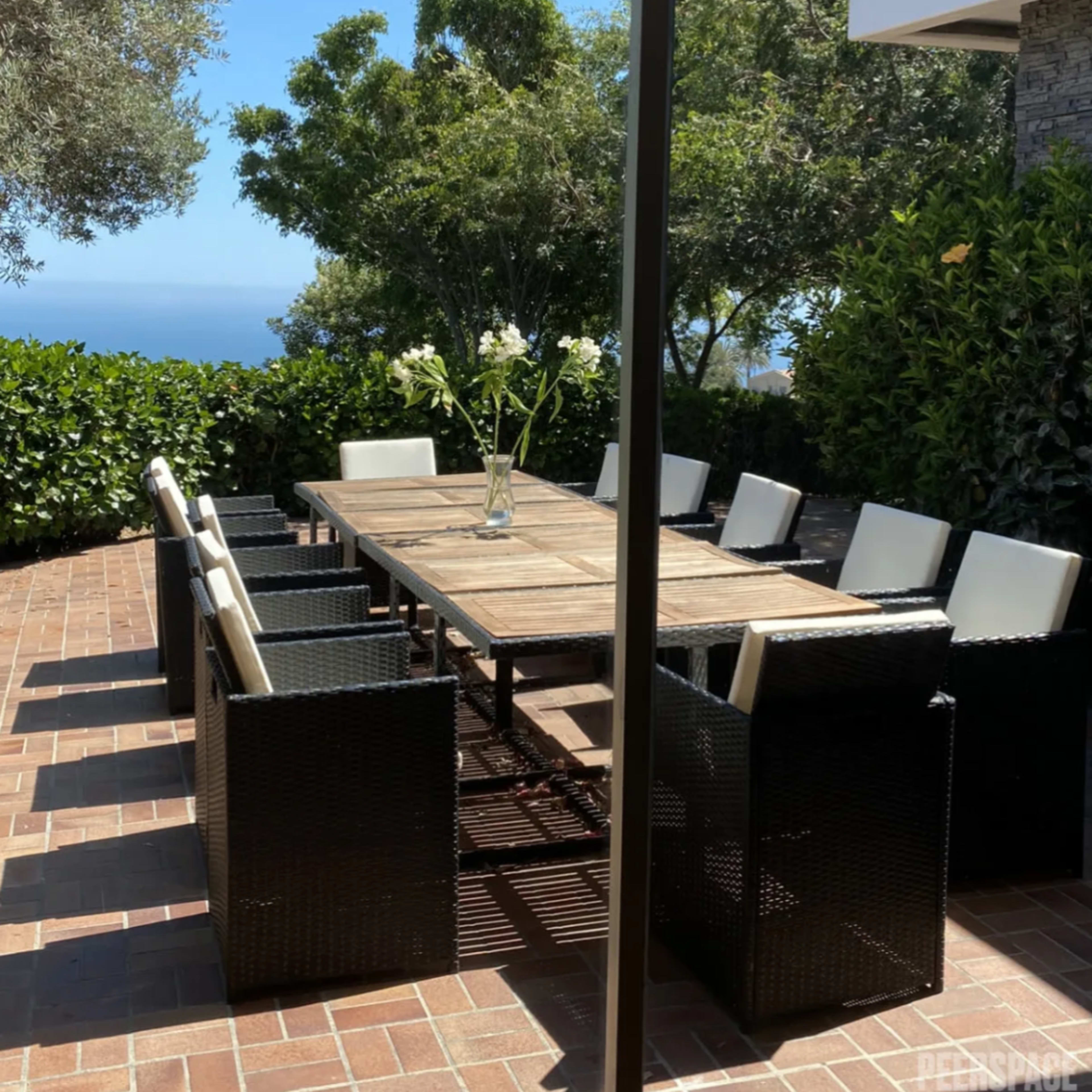 A wooden dining table surrounded by black wicker chairs sits in a landscaped outdoor area with views of greenery and the ocean.