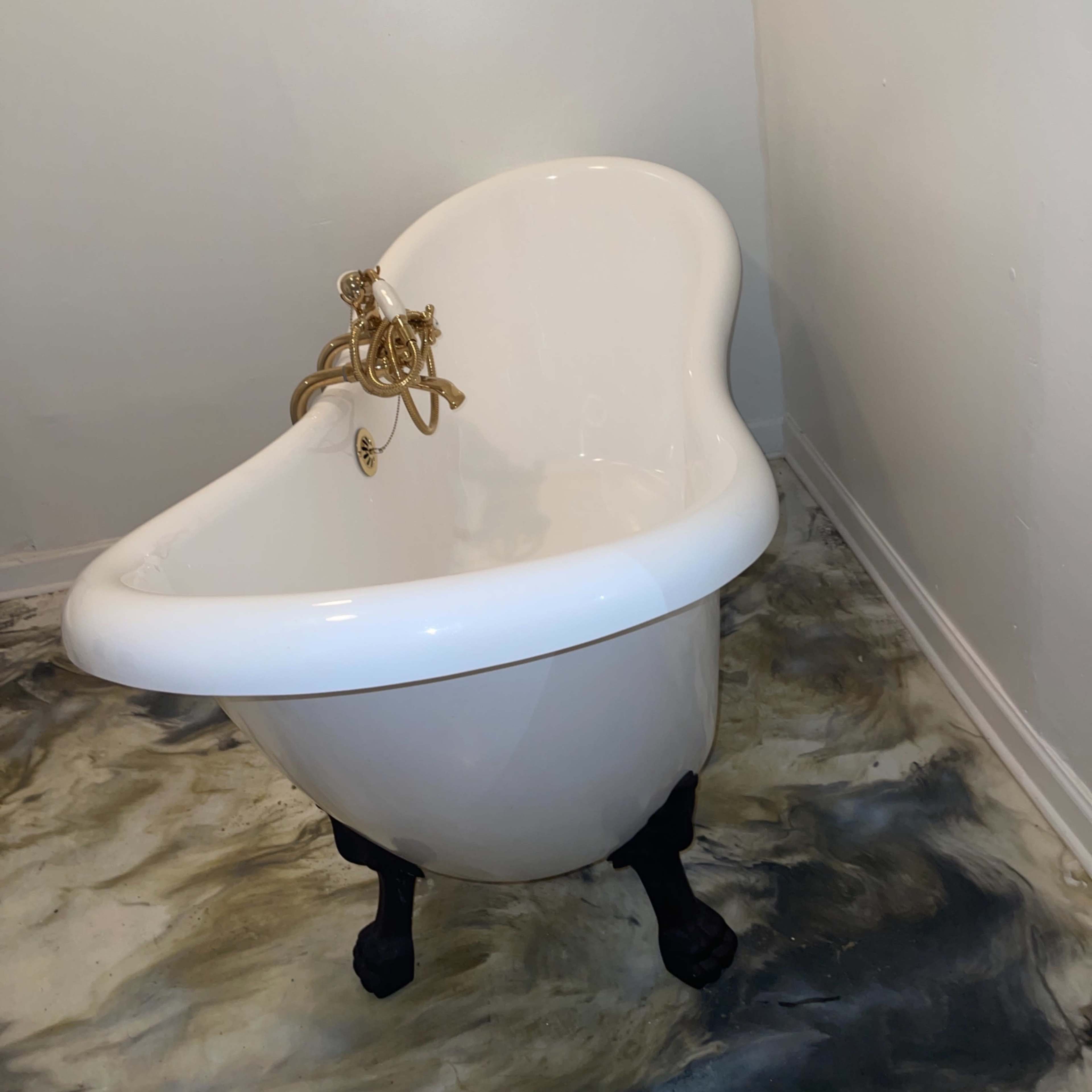 A white clawfoot bathtub with gold fixtures sits in the corner of a bathroom with a marbled floor.