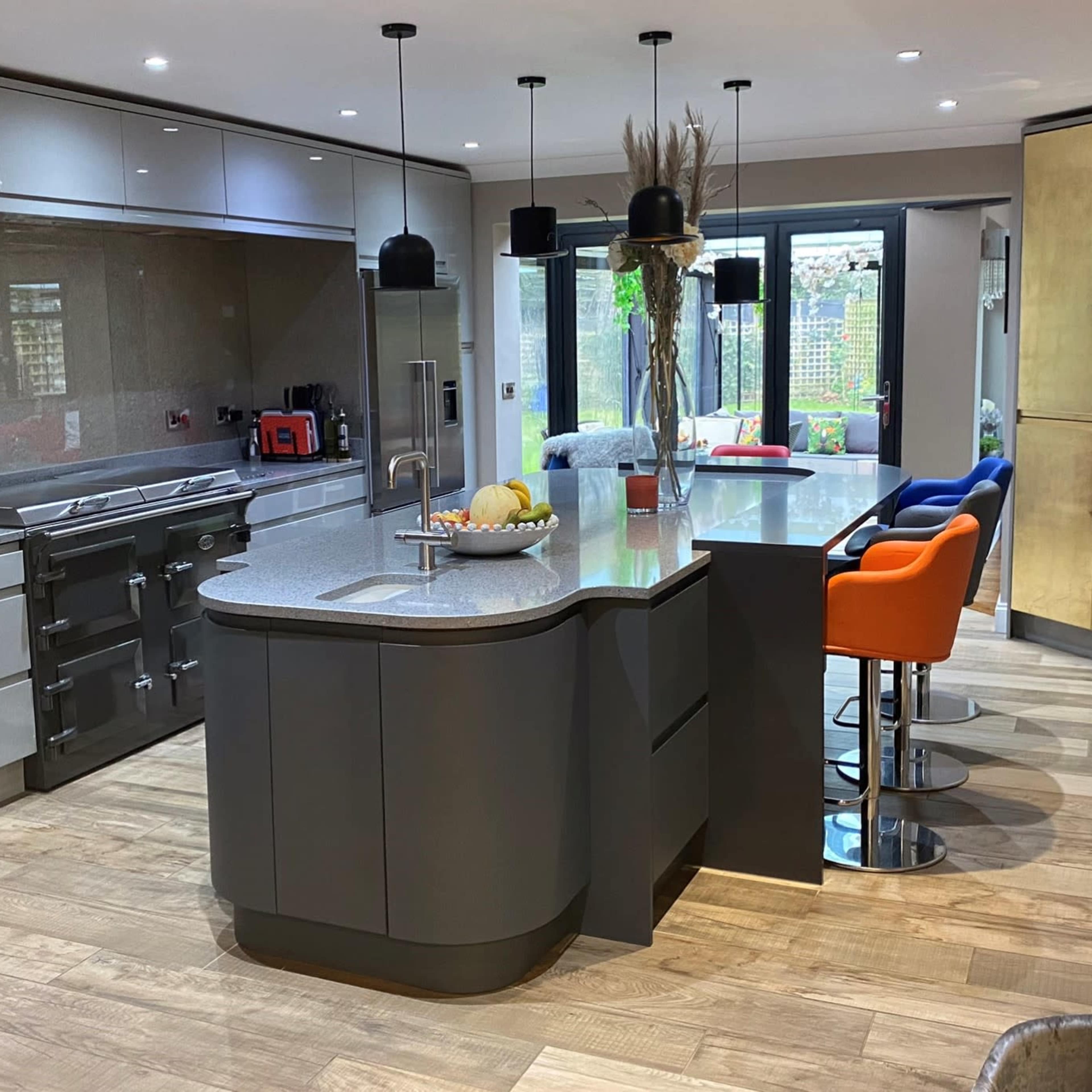 A modern kitchen features a central island with colorful bar stools, sleek cabinetry, and large windows overlooking a garden.
