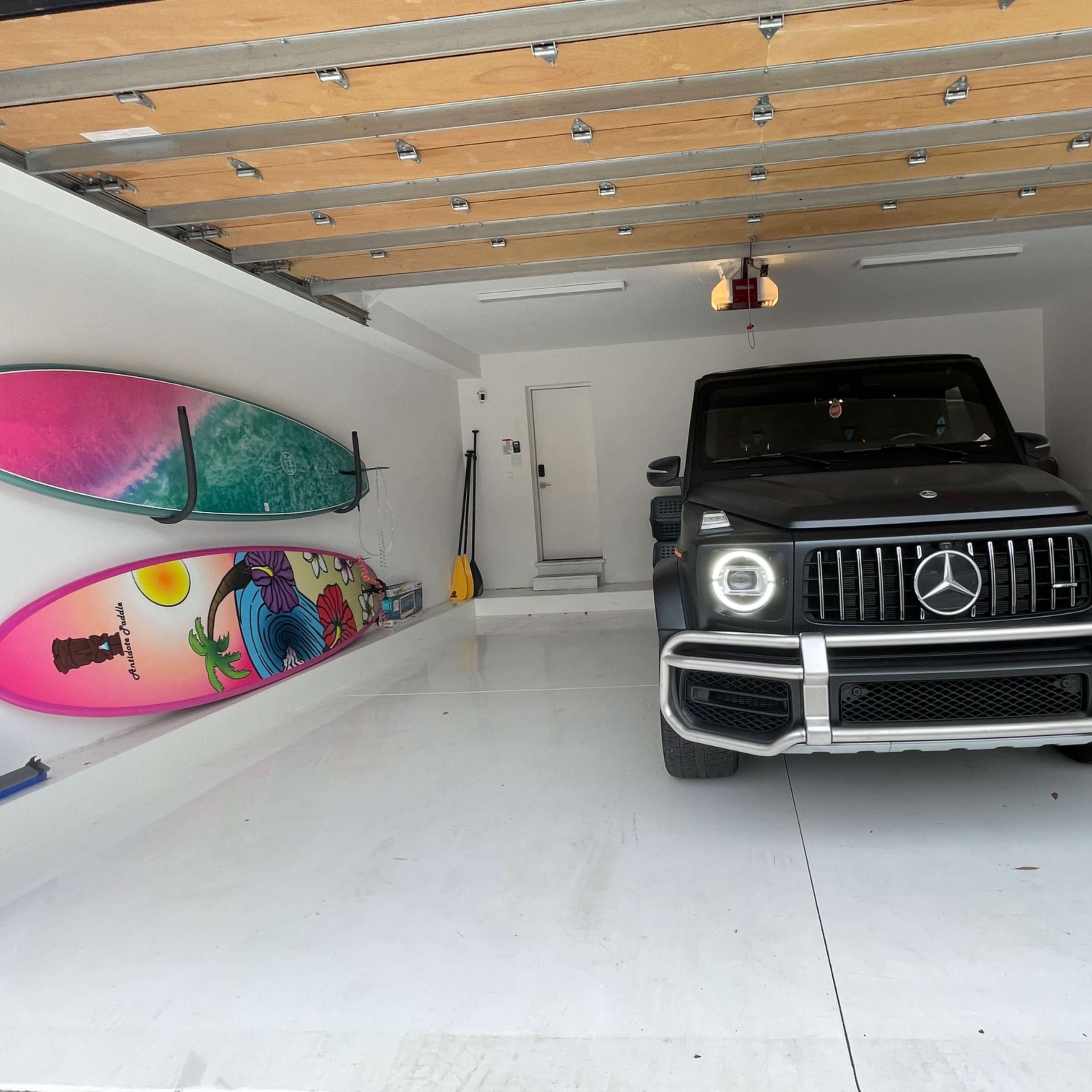 A black Mercedes-Benz G-Class is parked in a garage alongside two colorful surfboards.