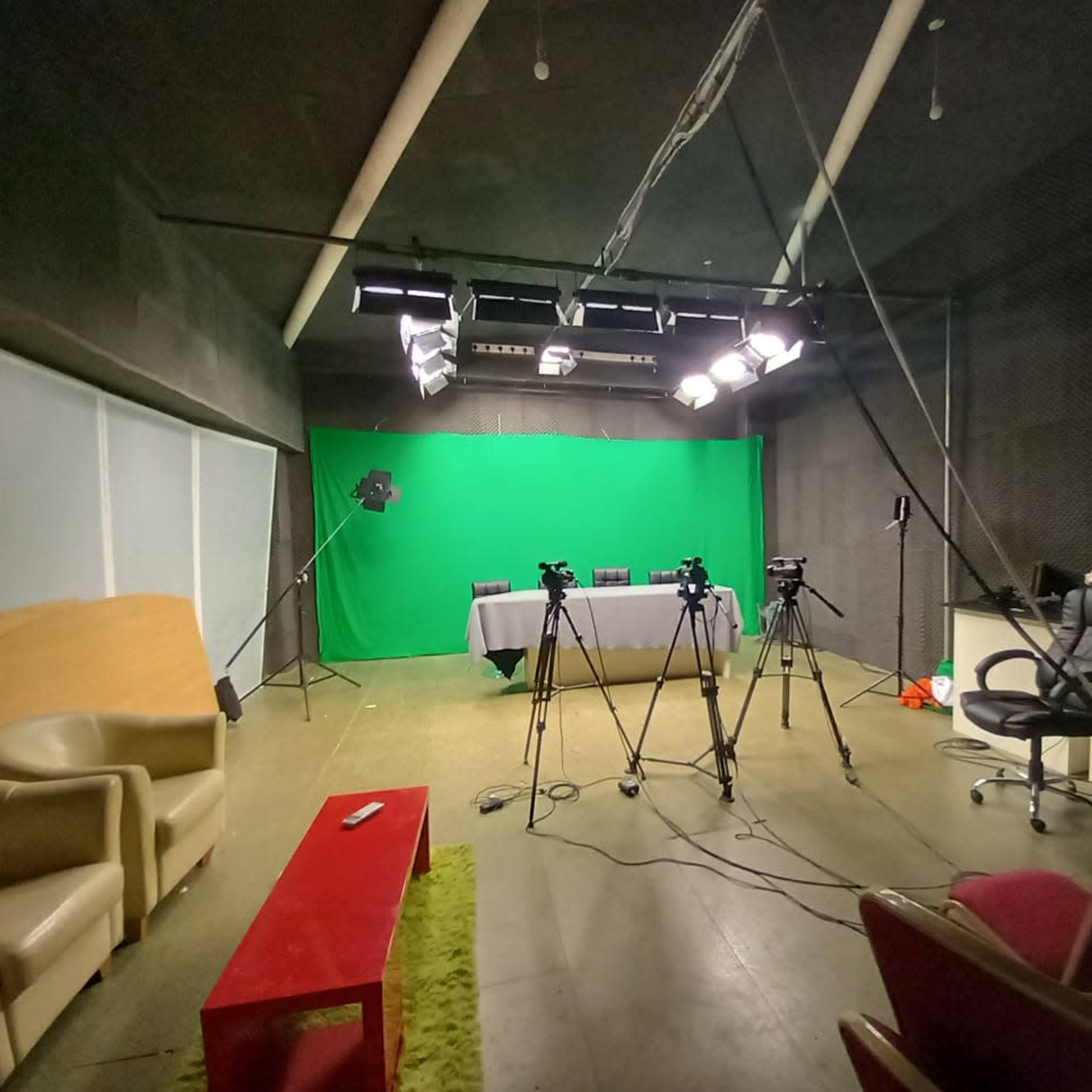 An empty studio with a green screen, several cameras on tripods, and a table set up in the center, along with chairs and lighting equipment.