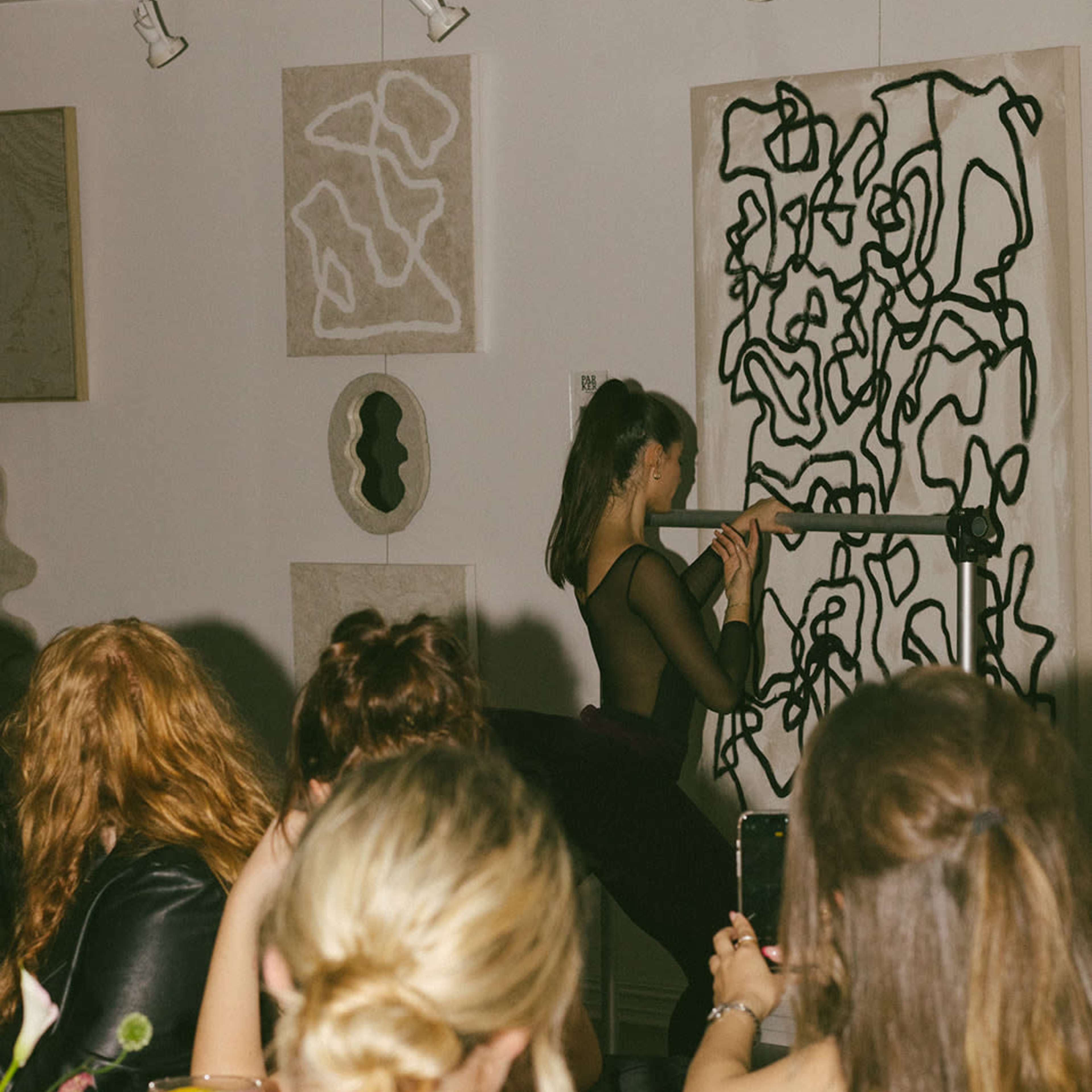 A performer plays a flute in front of an audience while abstract artworks hang on the walls of a gallery.