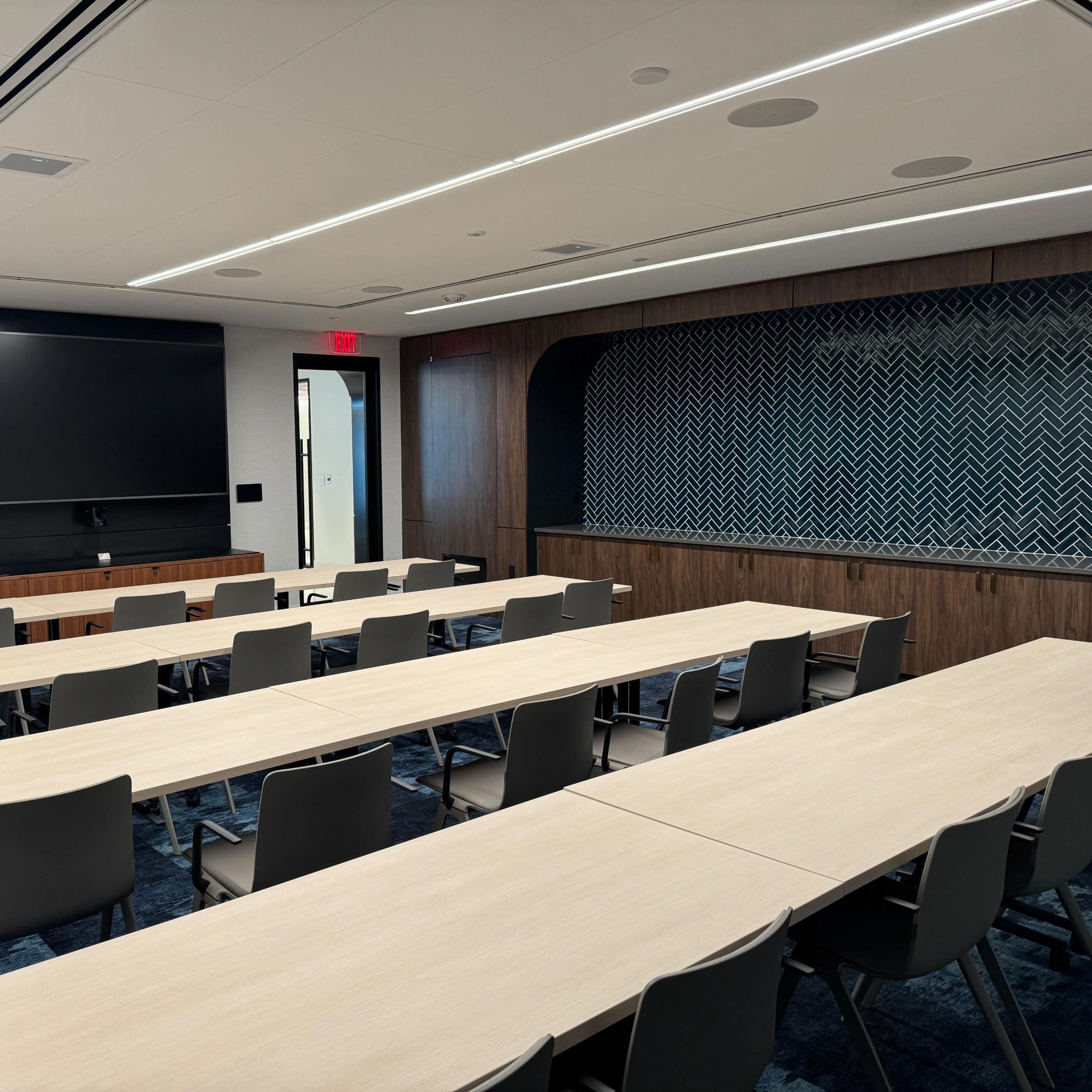 A modern conference room set up with multiple long tables and chairs, a large screen on the wall, and a podium in the corner.