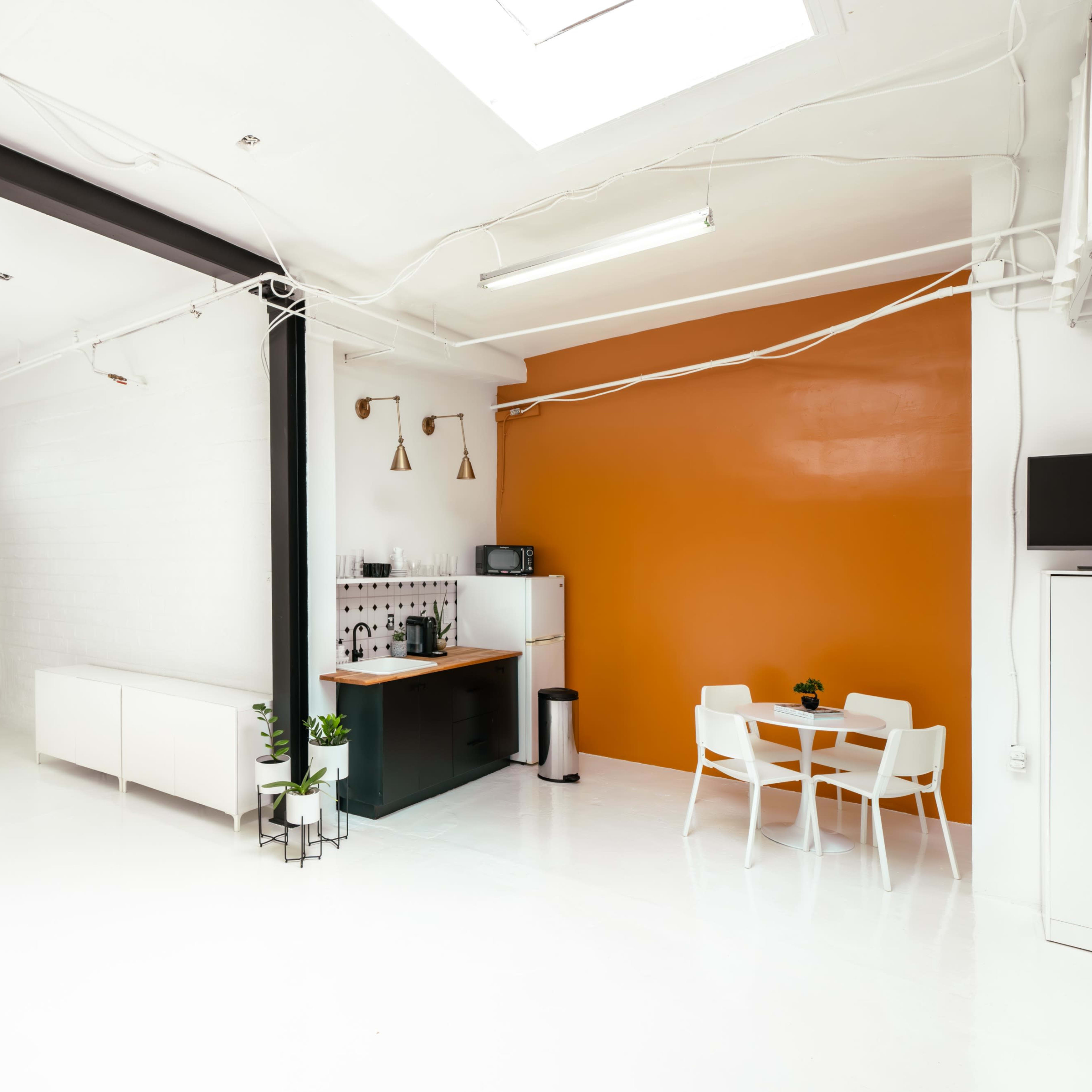 A spacious, minimalist interior with a kitchenette, a white table and chairs, and an orange accent wall.