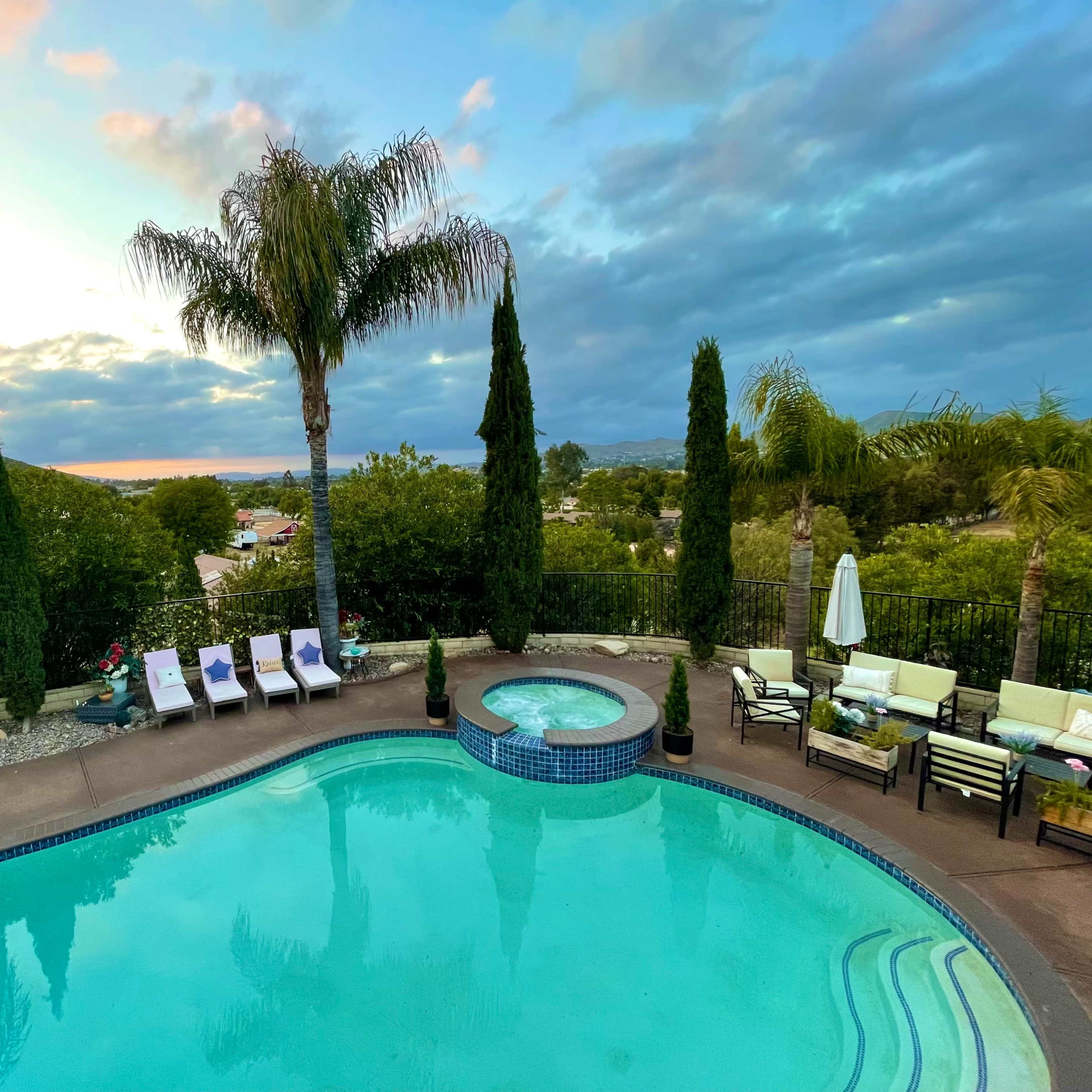 A swimming pool surrounded by palm trees and lounge chairs, with a hot tub nearby, set against a backdrop of a cloudy sky and distant hills.