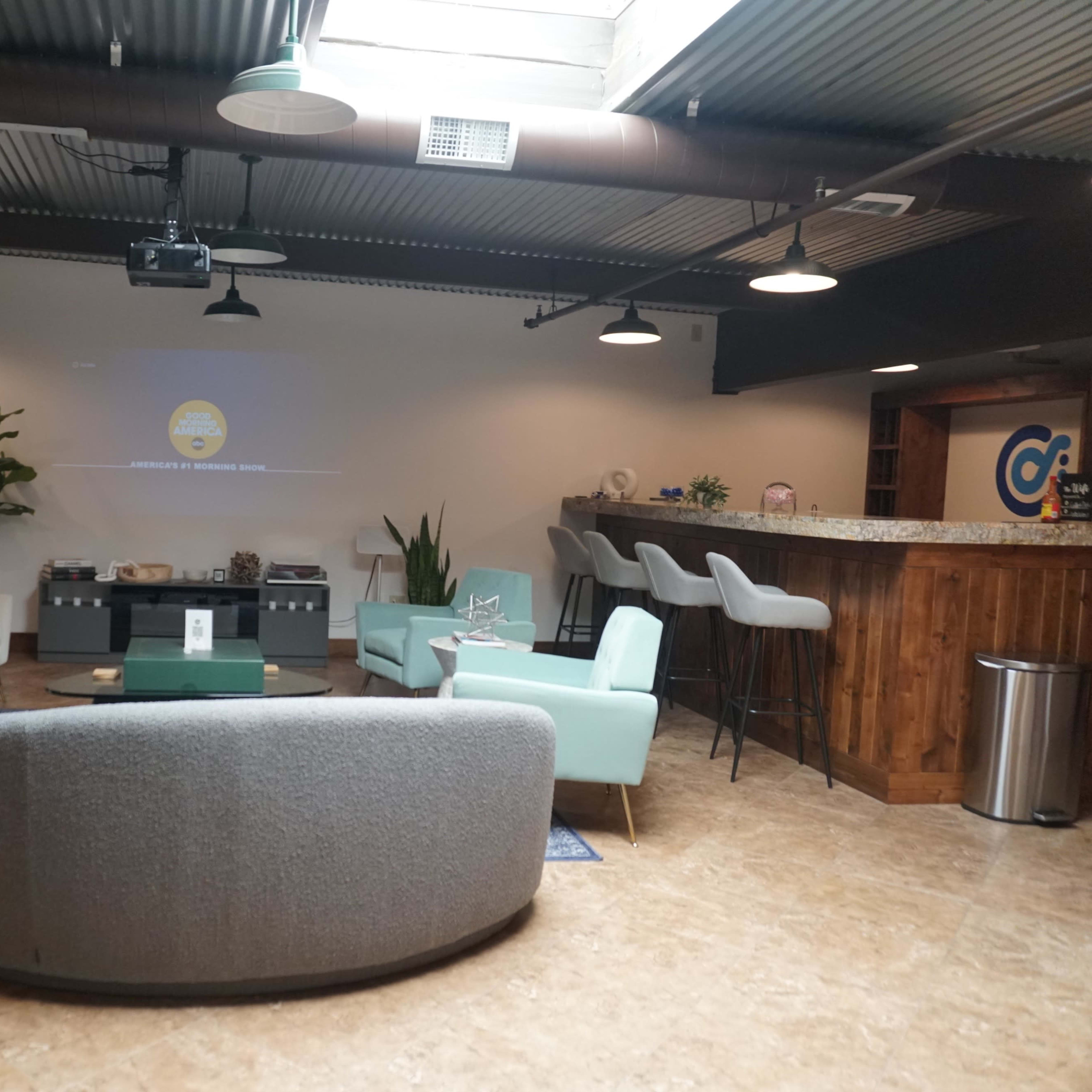 A modern lounge area with two light blue chairs, a grey sofa, a bar counter, and seating around a central coffee table under industrial-style lighting.