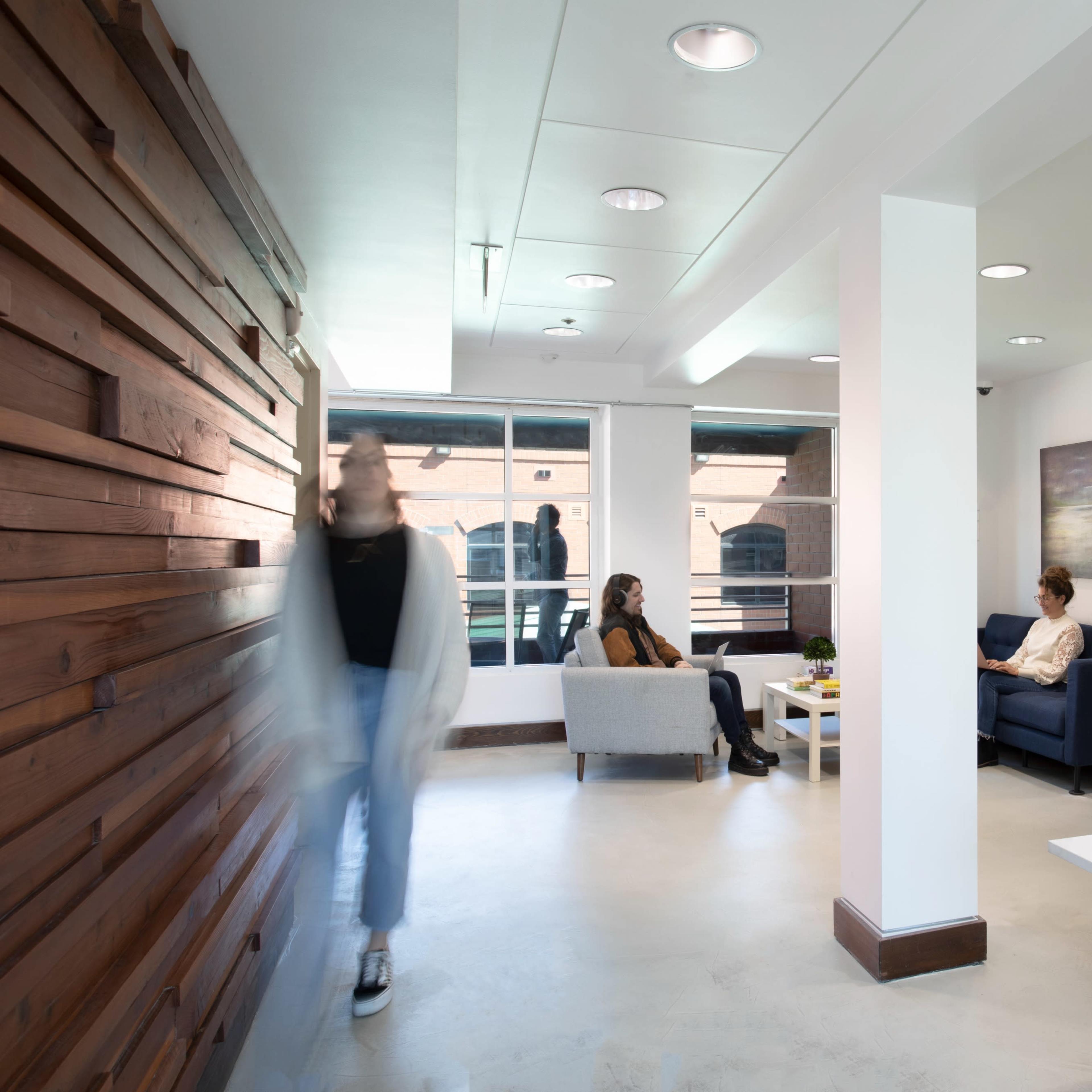 A modern office space features a wooden accent wall, a green plant wall, and individuals seated and interacting in a relaxed setting.