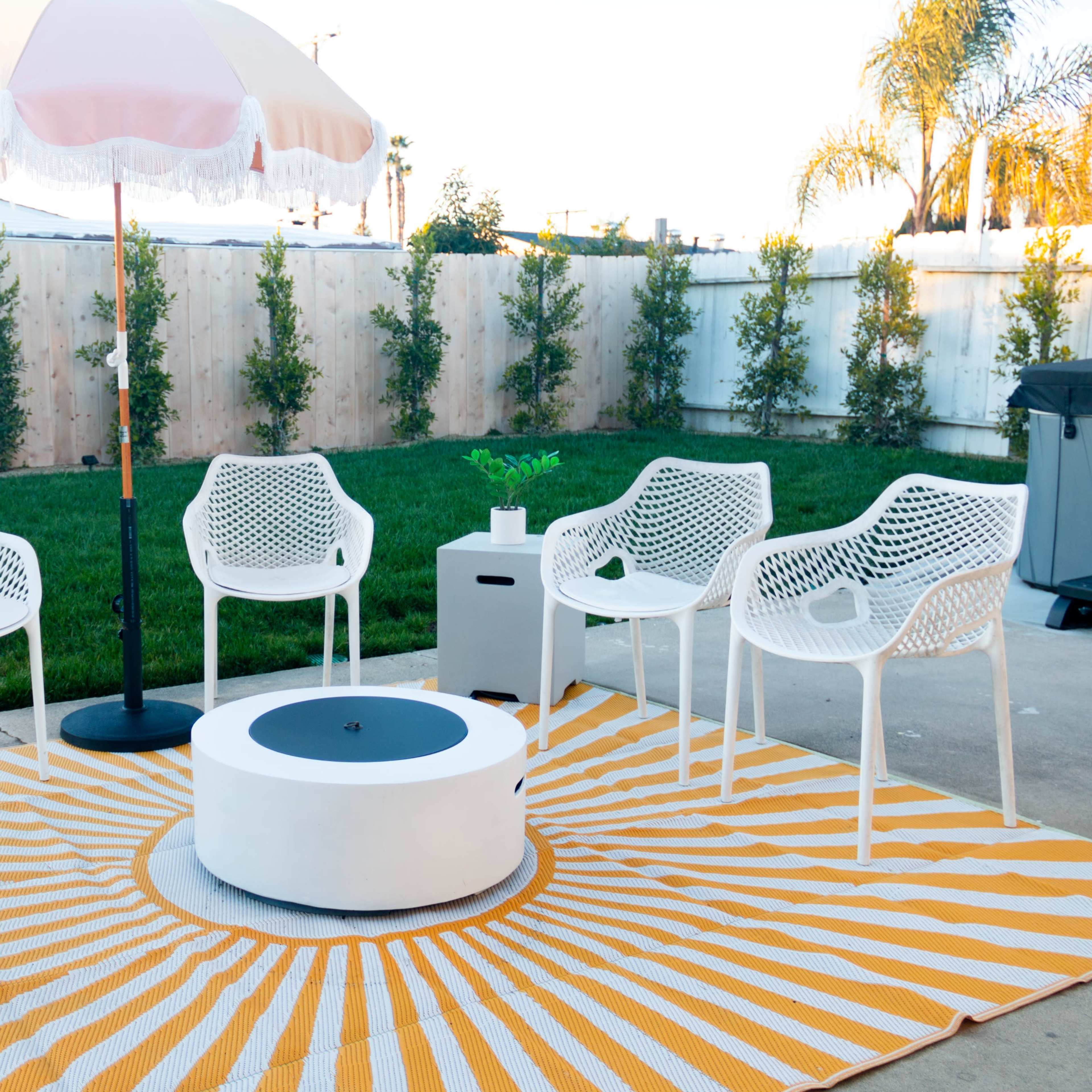 A circular outdoor seating area features six white chairs arranged around a central fire pit on a striped yellow and white rug, with a large umbrella providing shade.