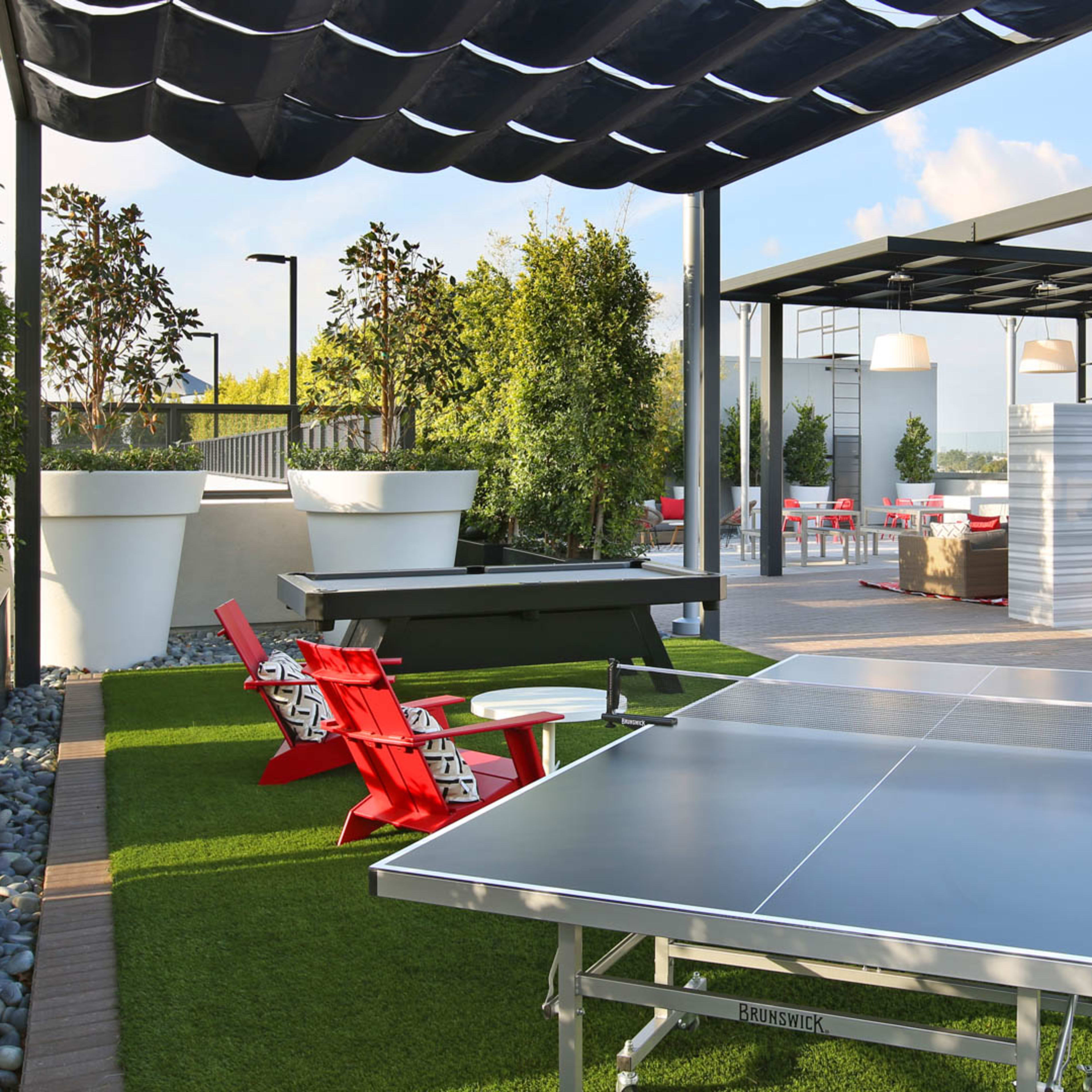 A recreational area featuring a ping pong table and red lounge chairs under a shaded pergola, surrounded by greenery and seating arrangements.