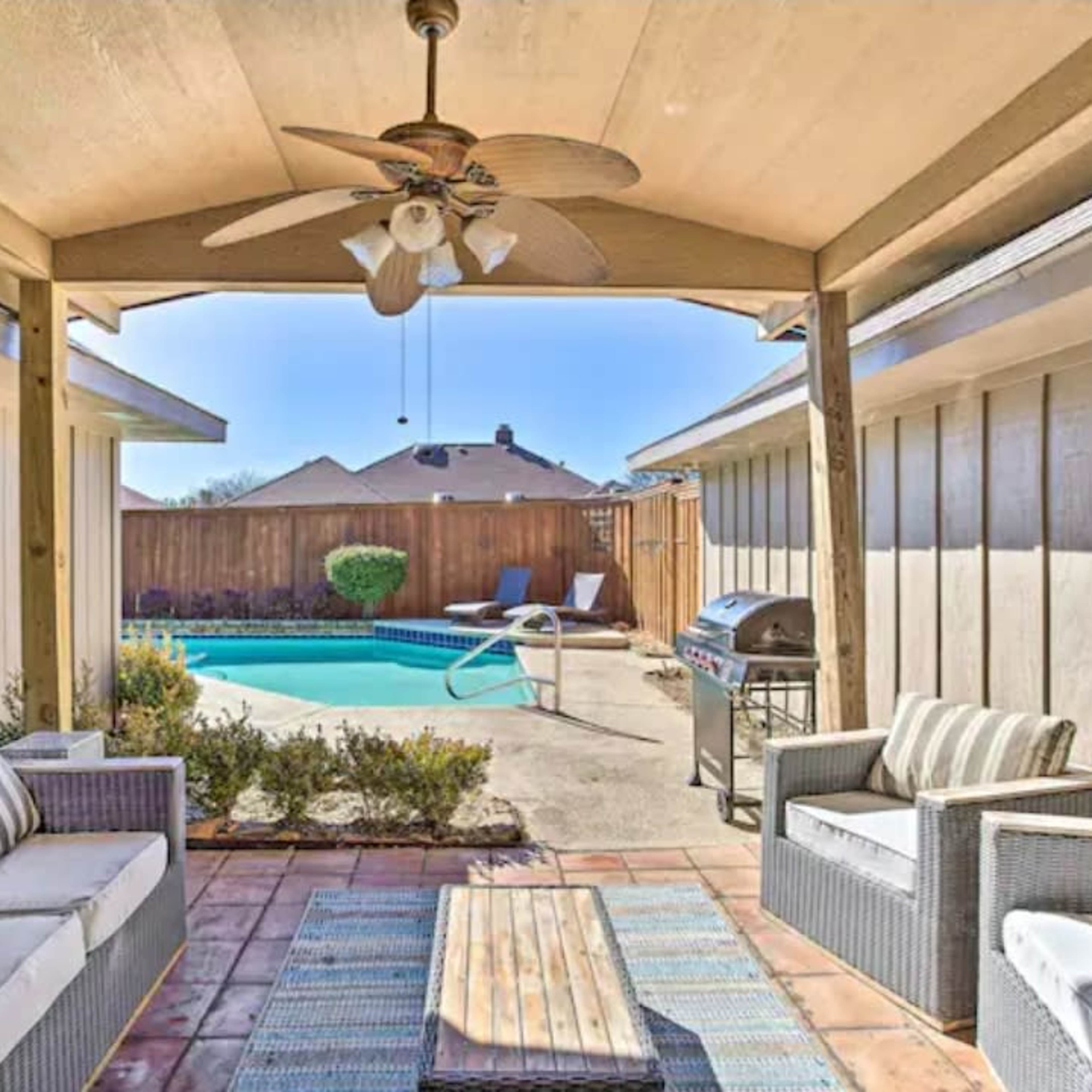 A covered patio area with seating and a coffee table, overlooking a swimming pool in a backyard.