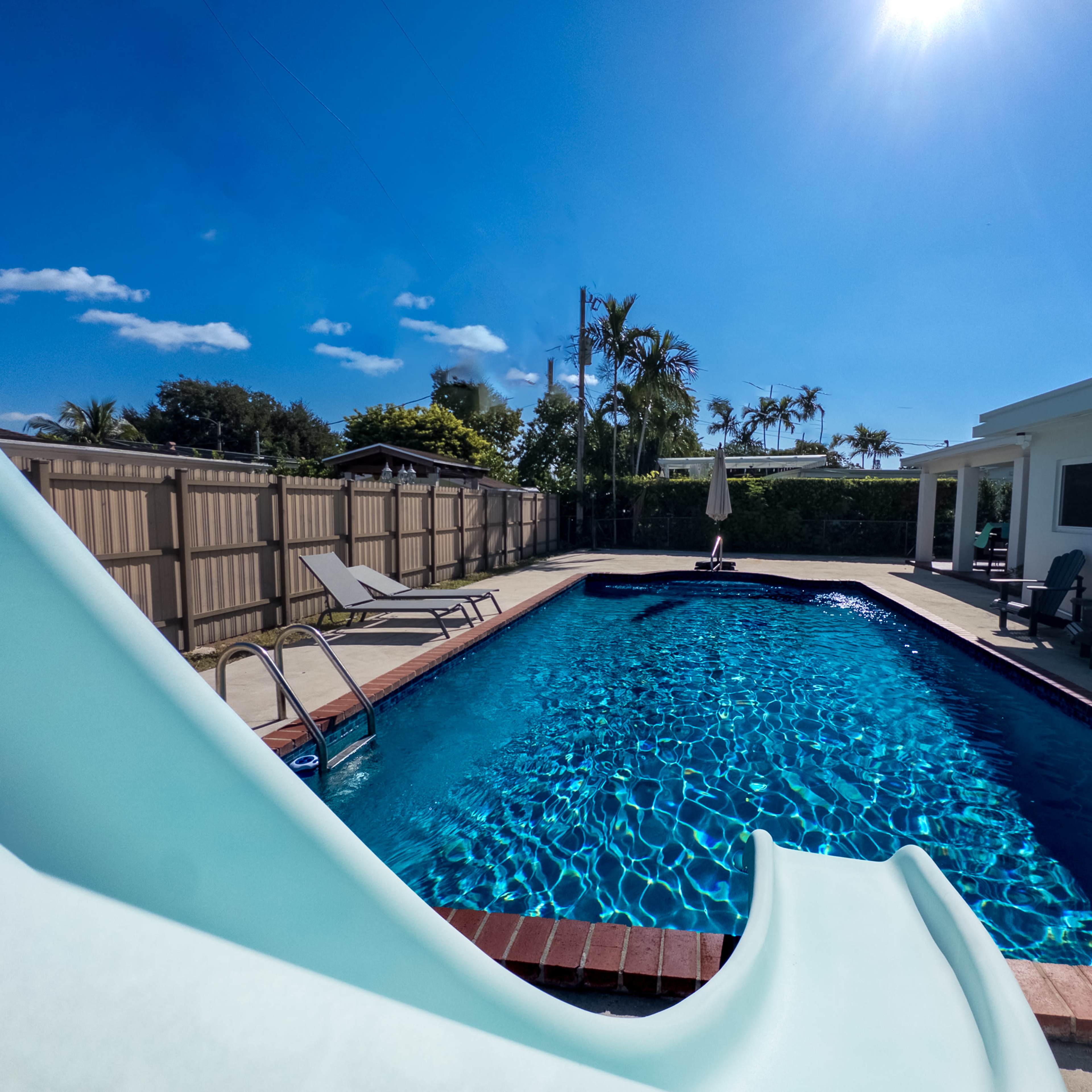 A clear swimming pool with a slide, surrounded by lounge chairs and a house under a bright blue sky.