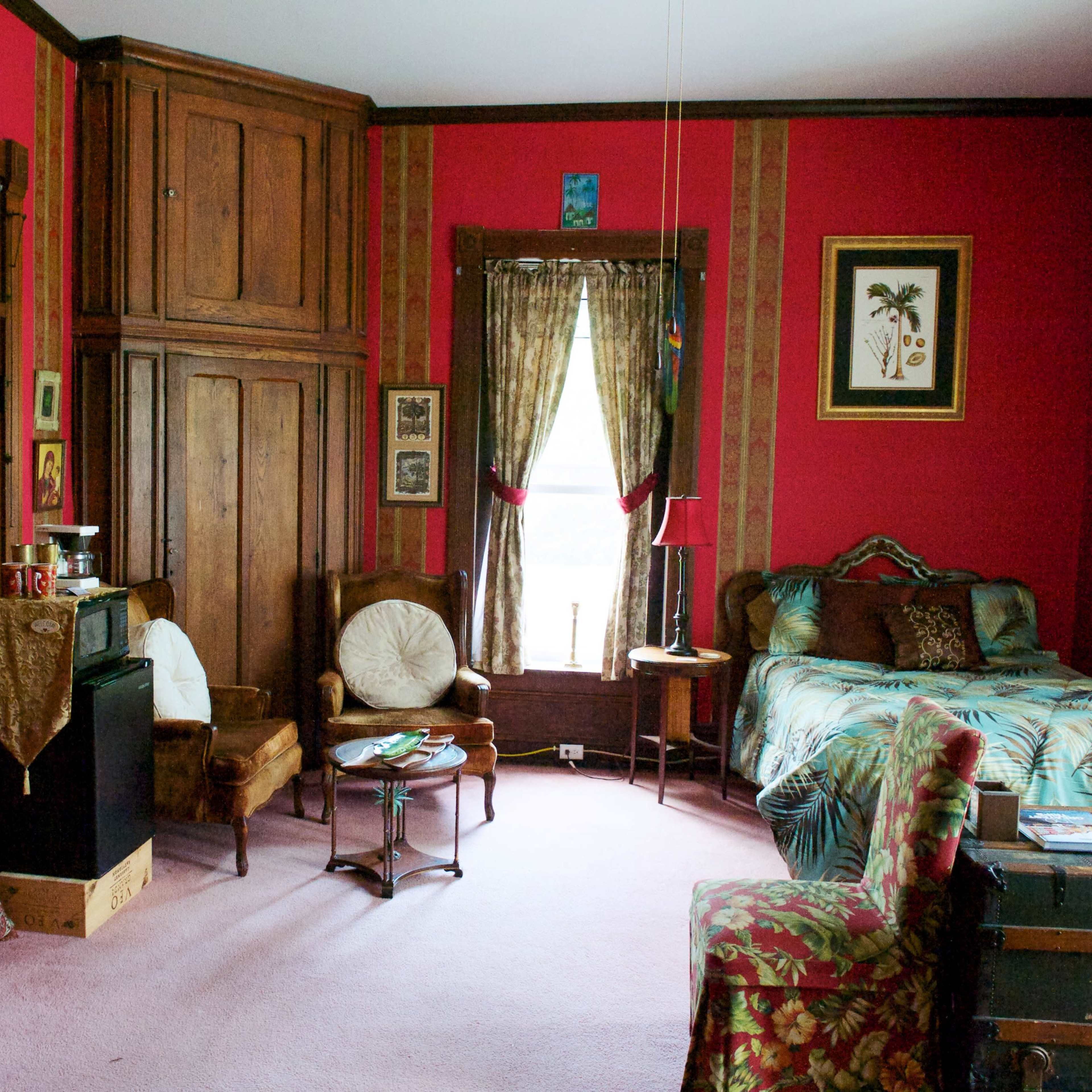 A cozy bedroom with red walls, a bed with patterned bedding, an antique dresser, and various pieces of wall art.