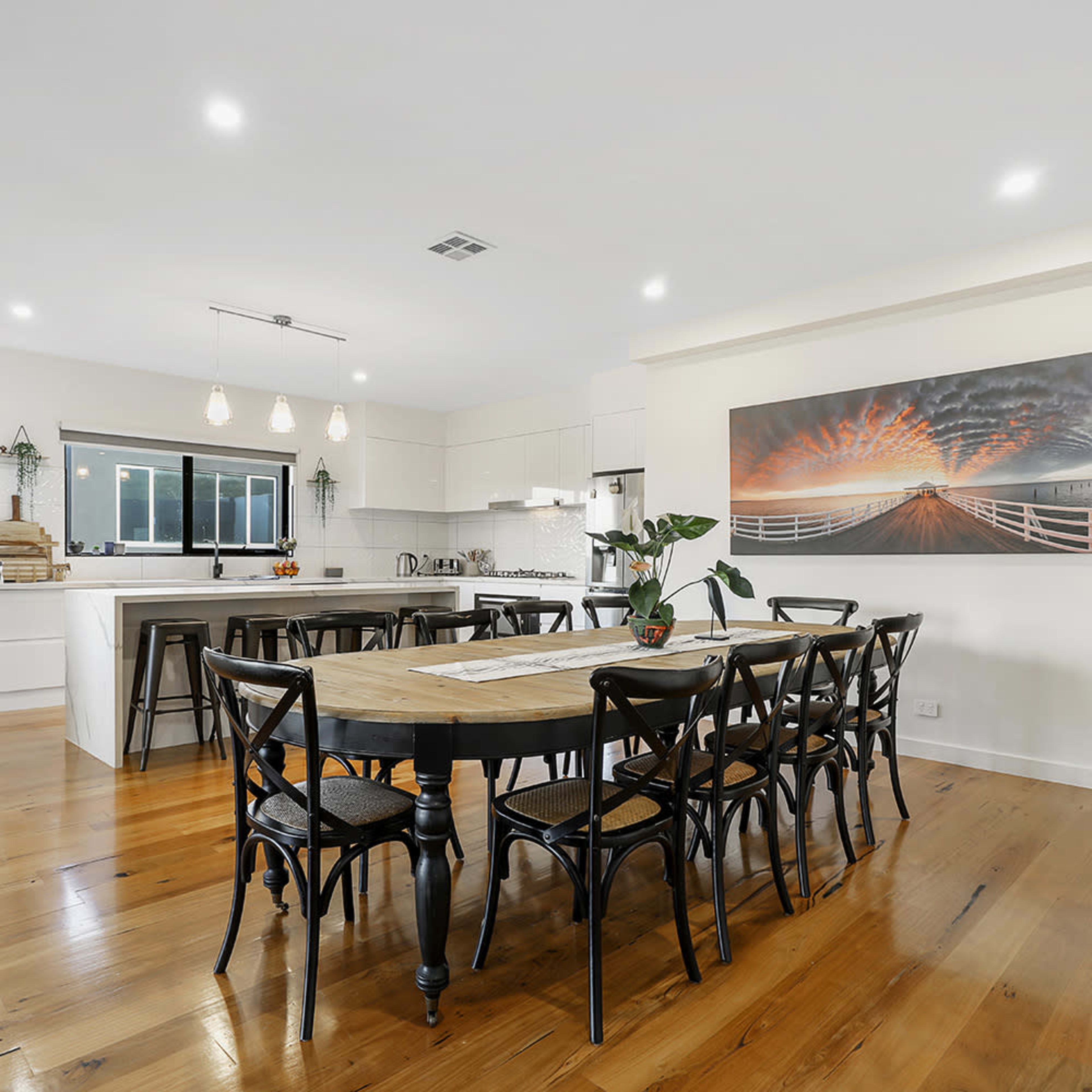 A spacious dining area features a large wooden table surrounded by black chairs, with modern cabinetry and a bright kitchen in the background.