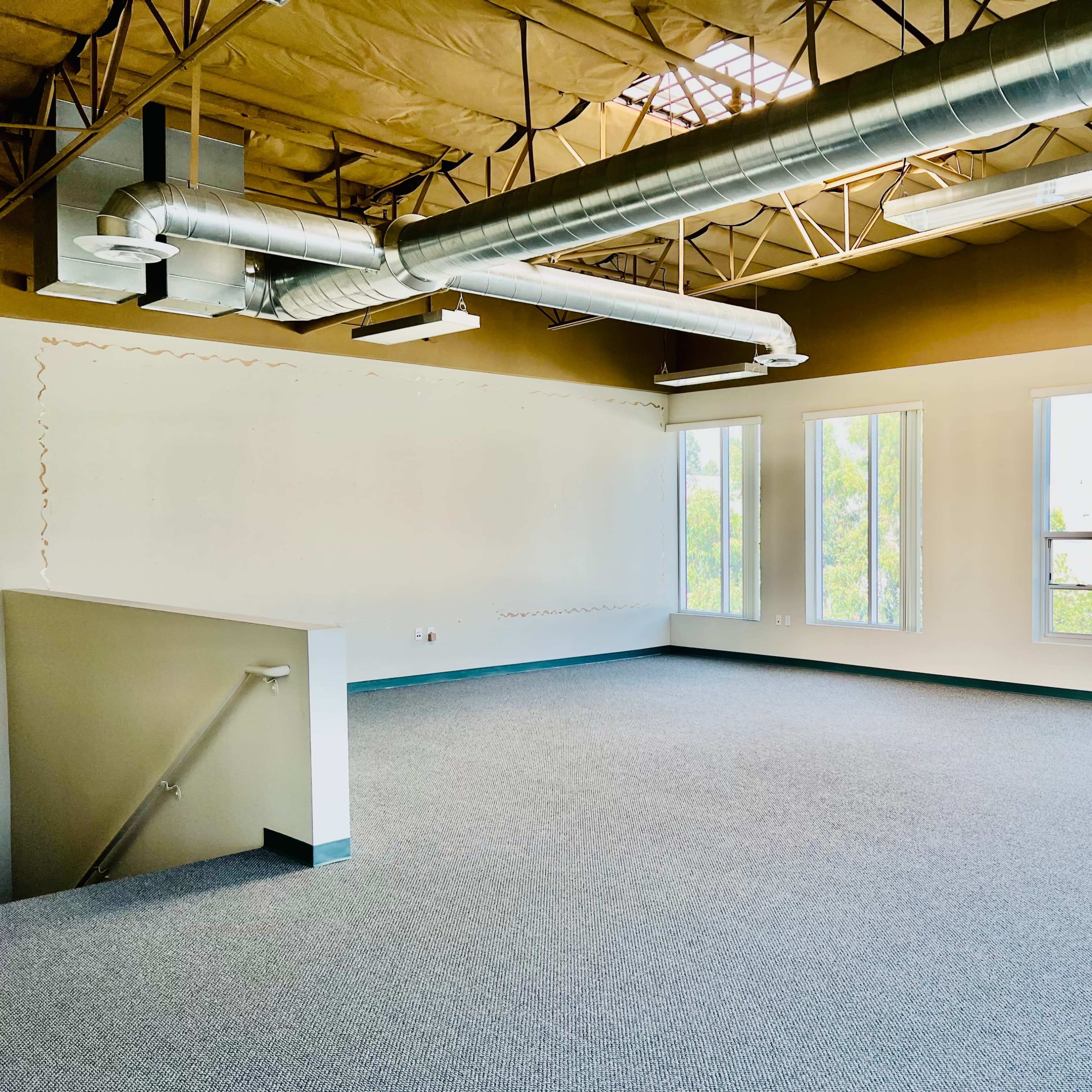 A spacious, empty room with large windows, a whiteboard, and exposed ventilation ducts above.