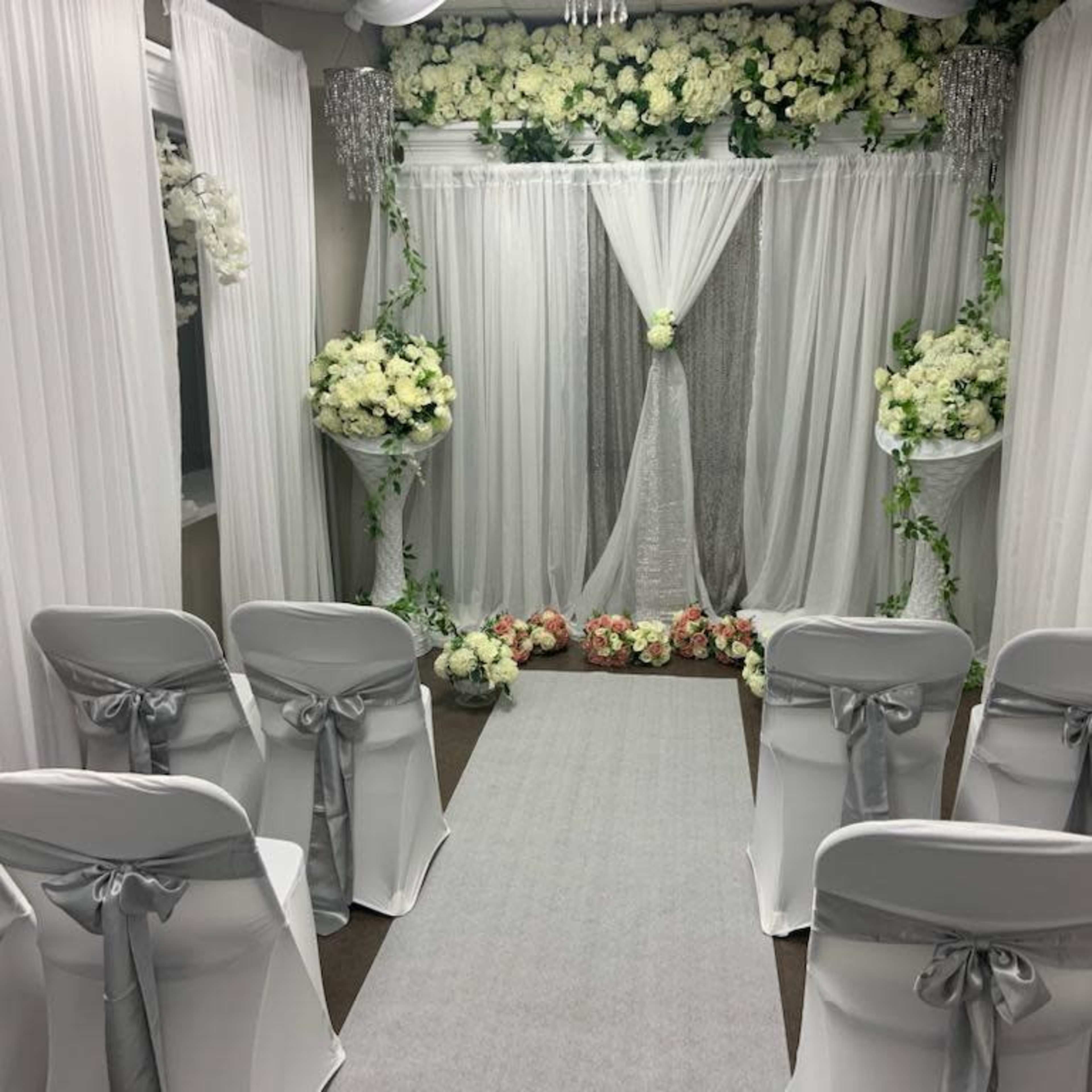A wedding ceremony setup with white drapery, floral arrangements, and chairs neatly aligned along a central aisle.