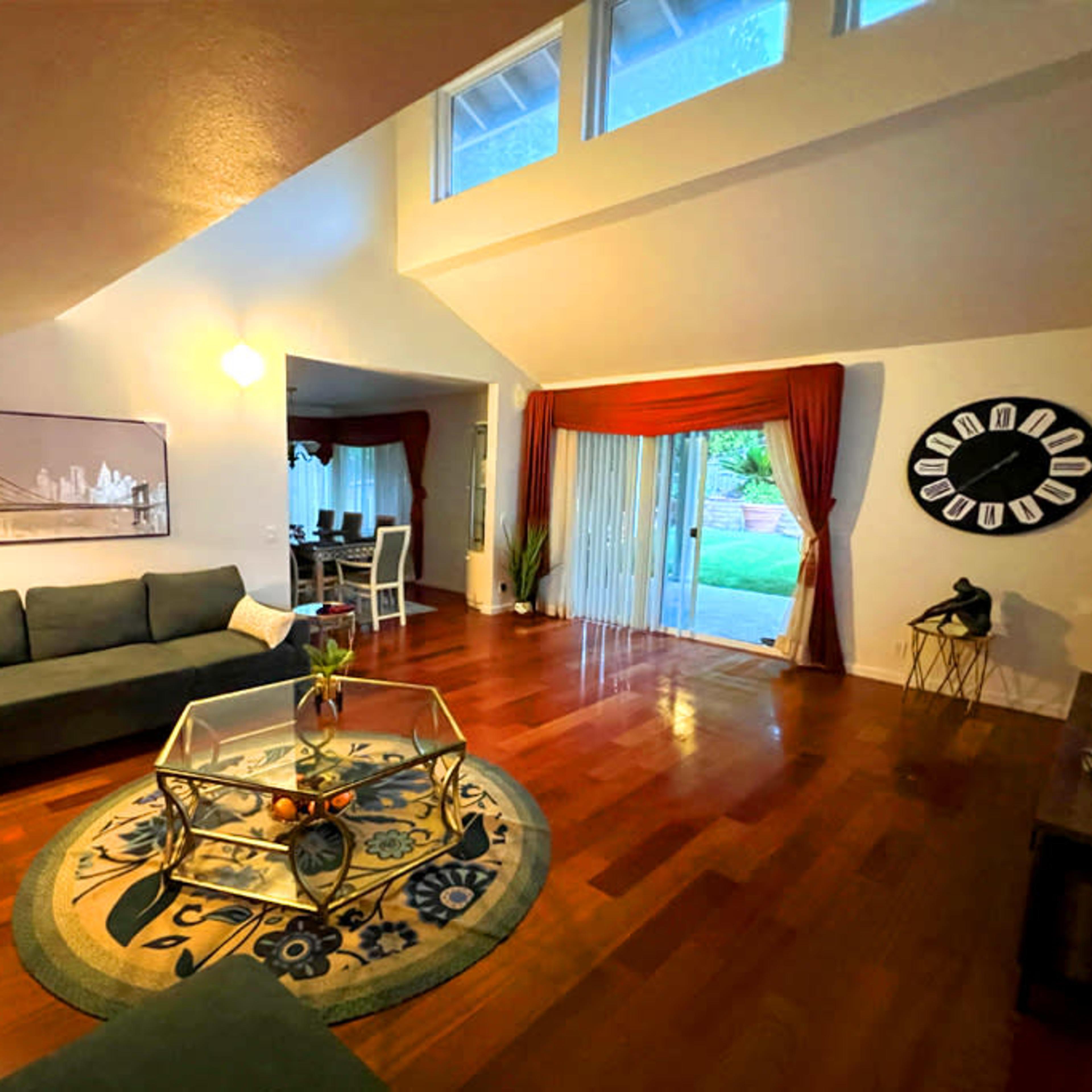A living room with a modern design featuring dark sofas, a glass coffee table, hardwood floors, and large windows opening to a backyard.