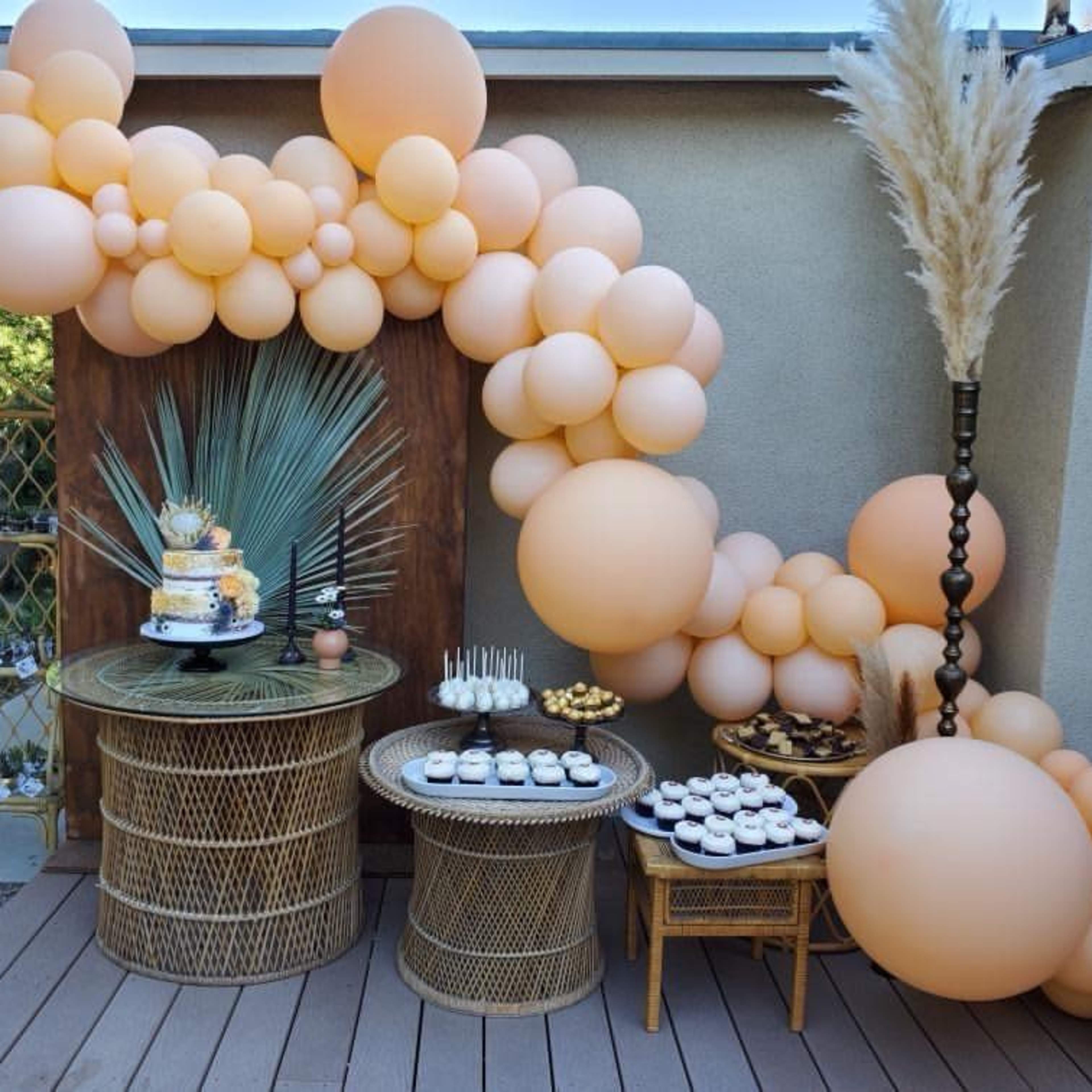 A decorated outdoor space featuring a large arrangement of peach-colored balloons, two woven tables with a tiered cake and assorted desserts.