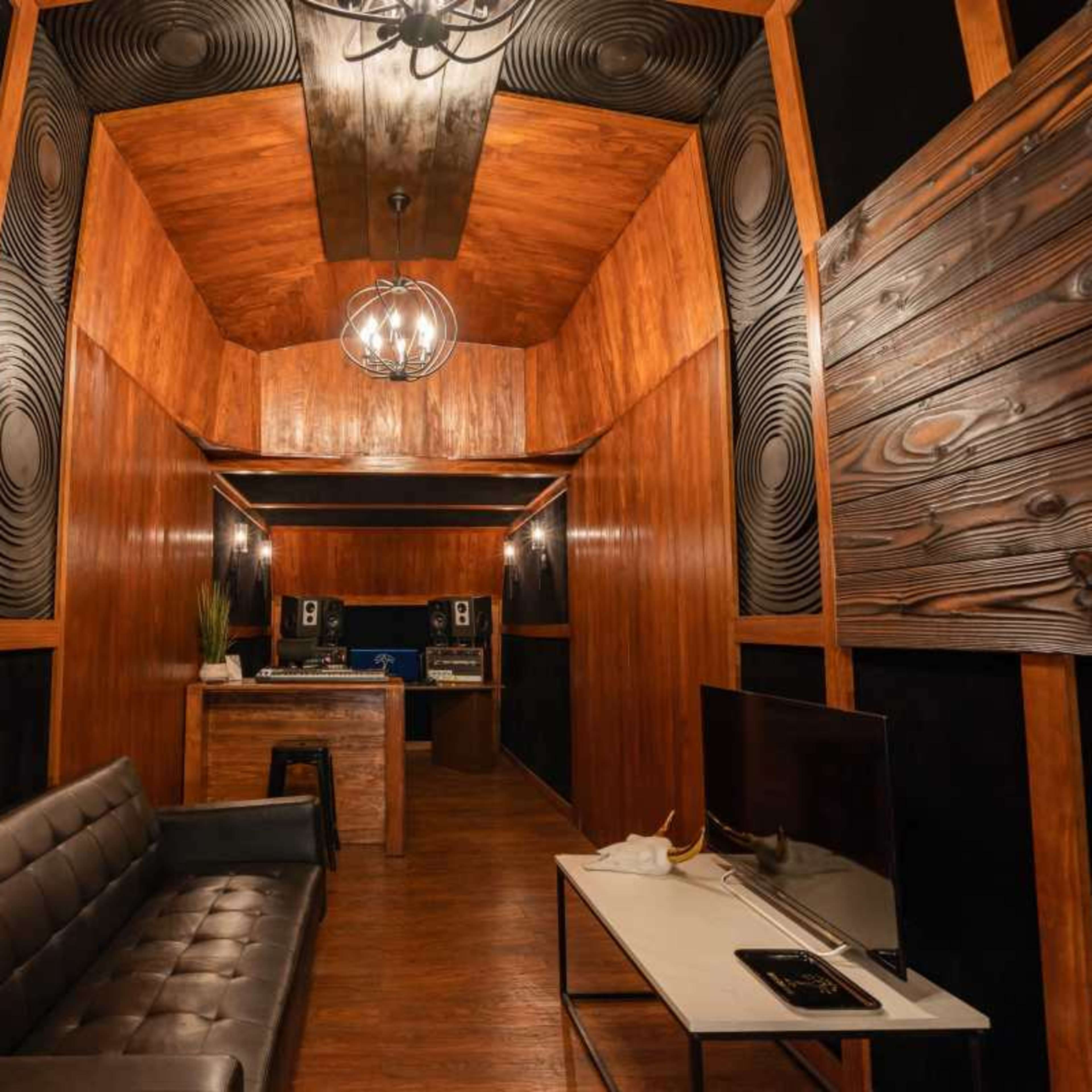 A music studio with wooden walls, a central seating area, and audio equipment visible in the background.