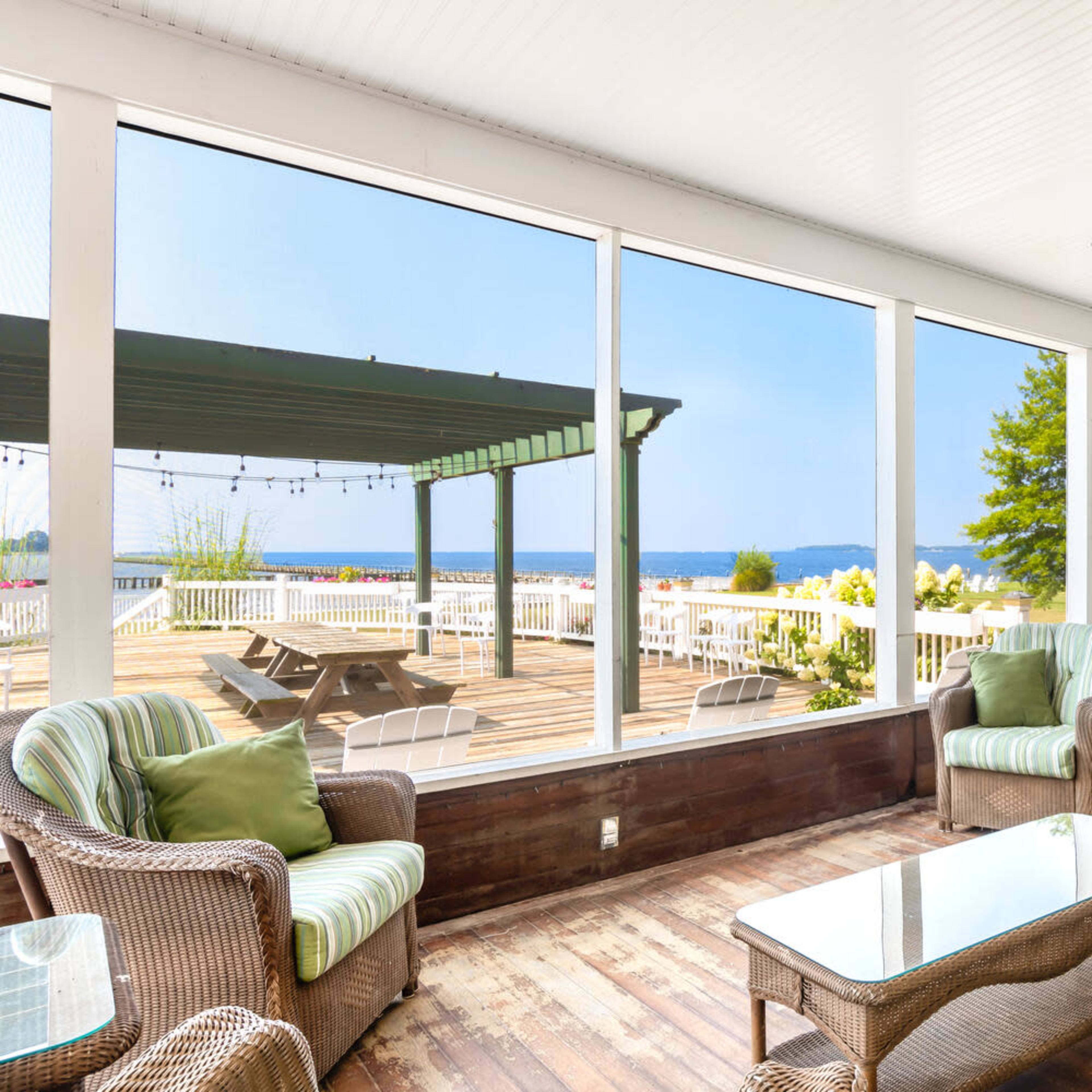 A screened-in porch features wicker furniture and a glass table, overlooking a deck and a view of a body of water and trees.