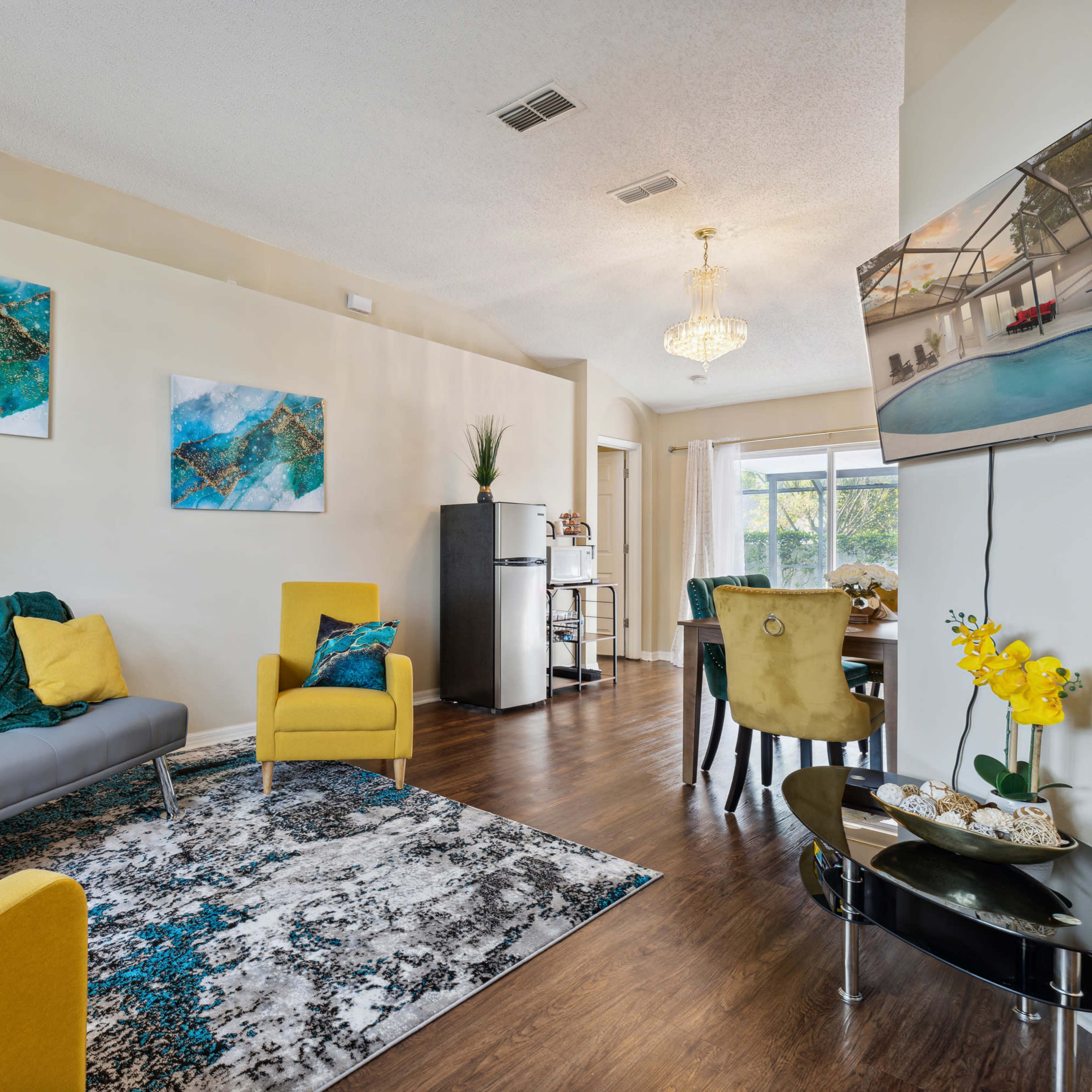 A modern living room features a gray sofa, yellow chairs, abstract artwork, and a dining area with a view of the outdoors.