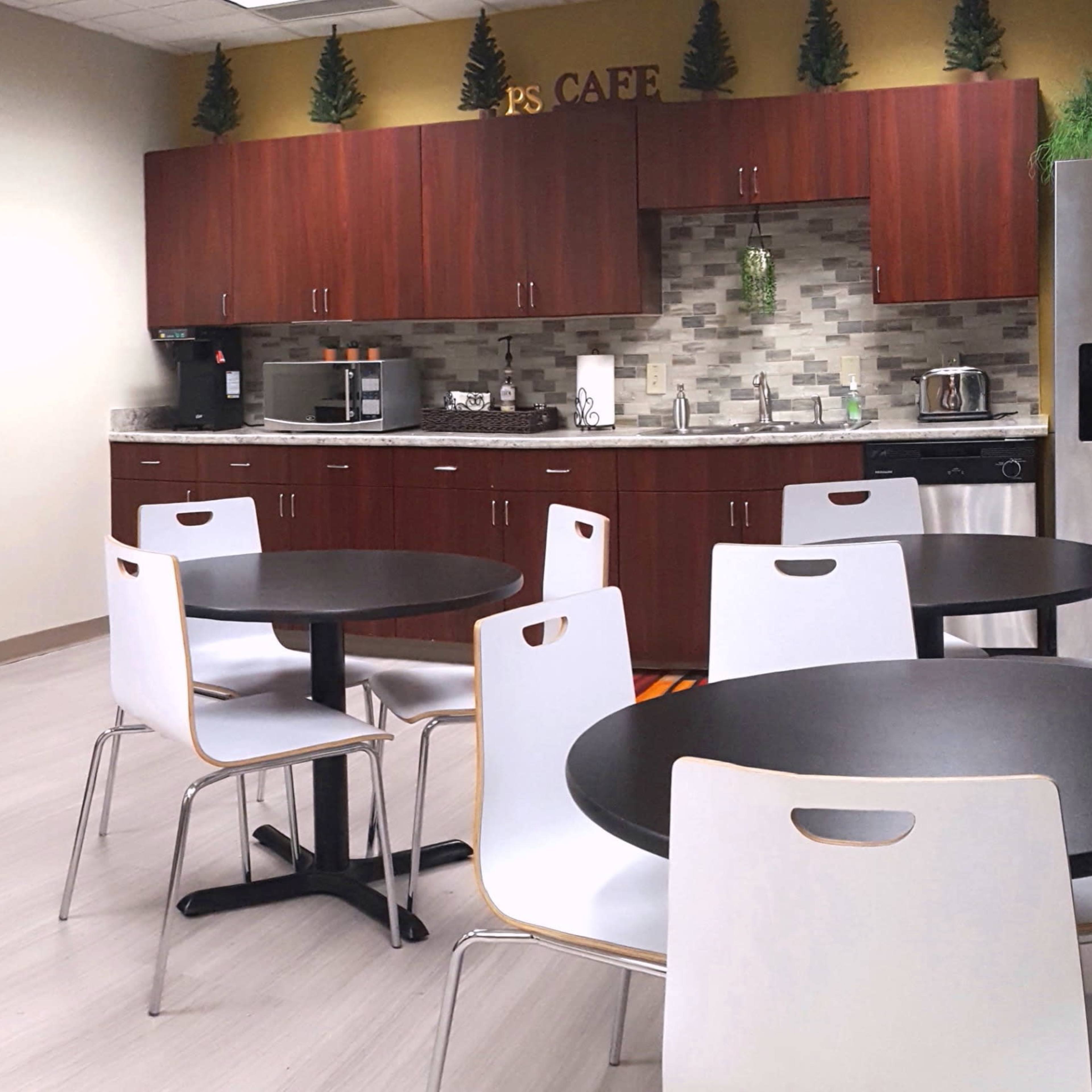 A modern breakroom with a kitchenette featuring brown cabinets, a coffee station, and several round tables surrounded by black and white chairs.