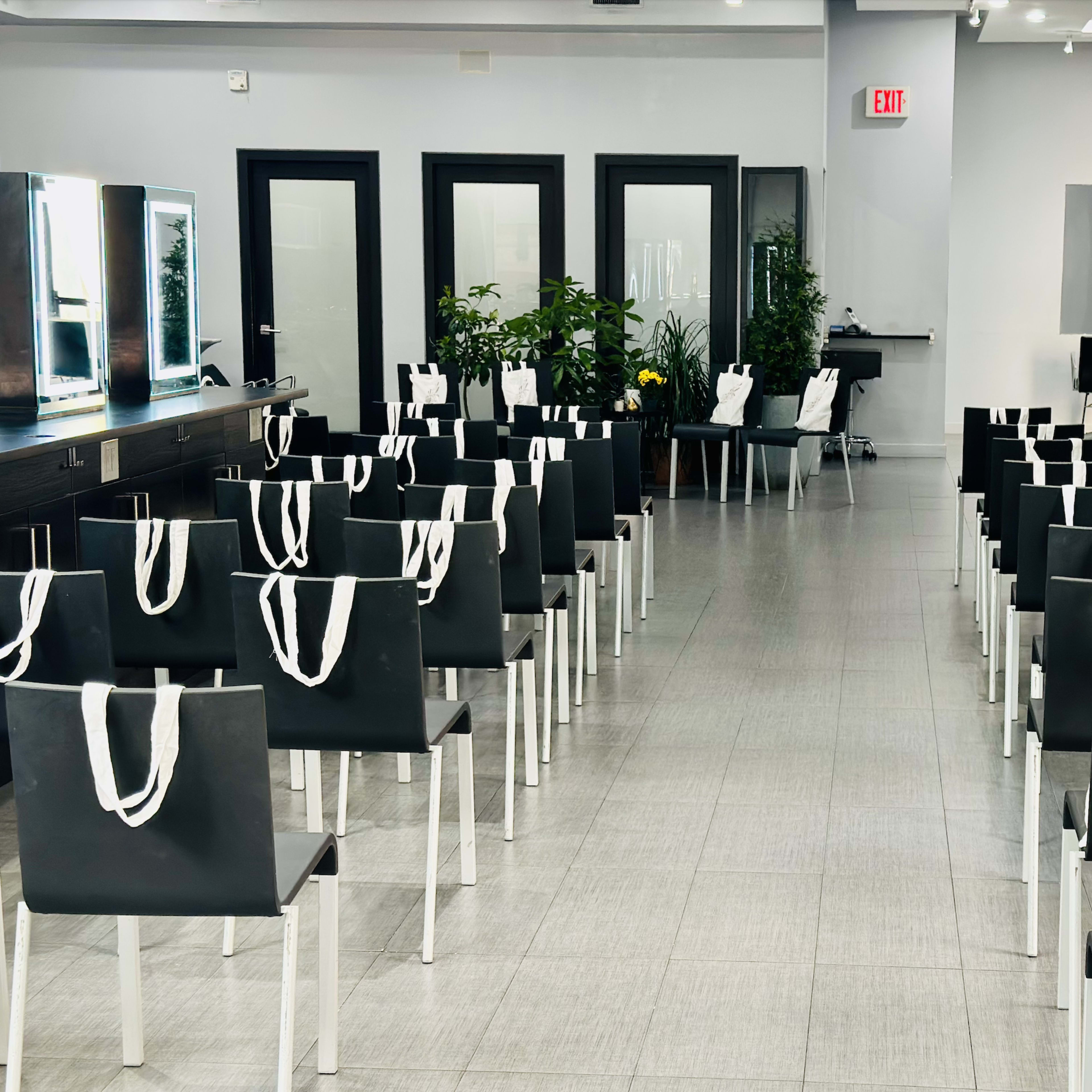 A row of empty black chairs with white tote bags placed on the backs, arranged in a well-lit salon or studio space.