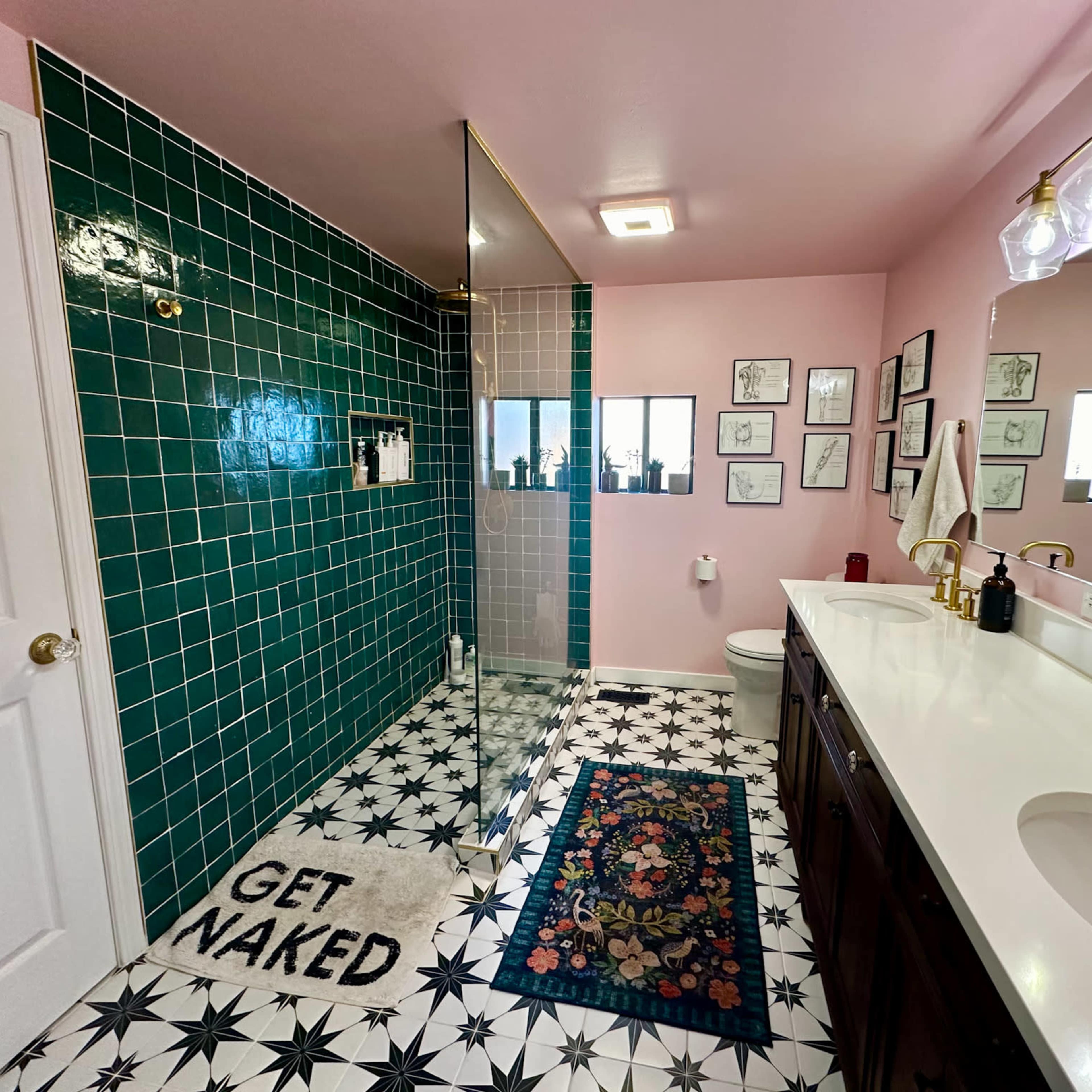 The bathroom features green tiled walls, a glass shower, a white countertop with dark wood cabinets, and a patterned floor with a decorative rug.