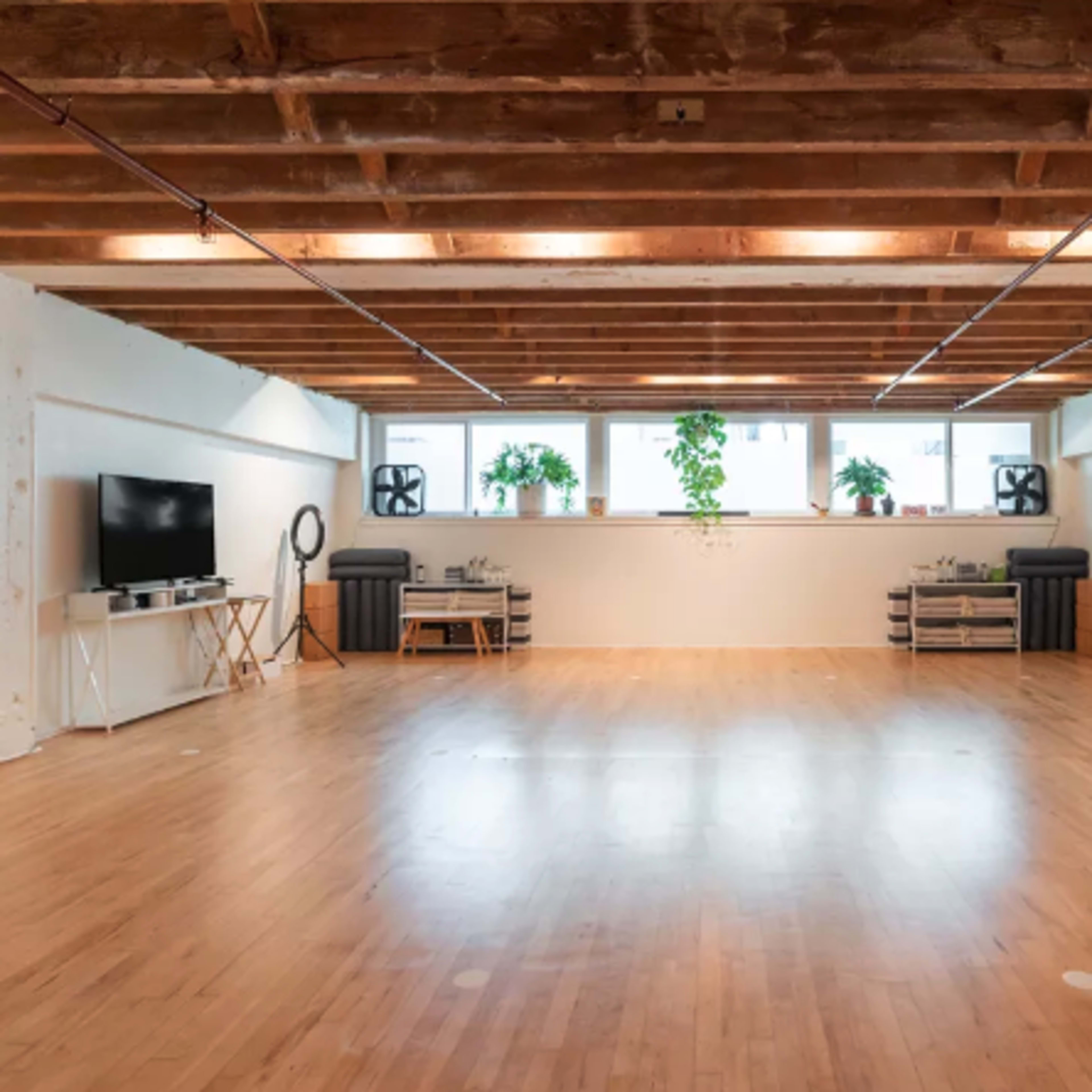 A spacious, empty room with hardwood flooring, exposed wooden beams, large windows, and potted plants.