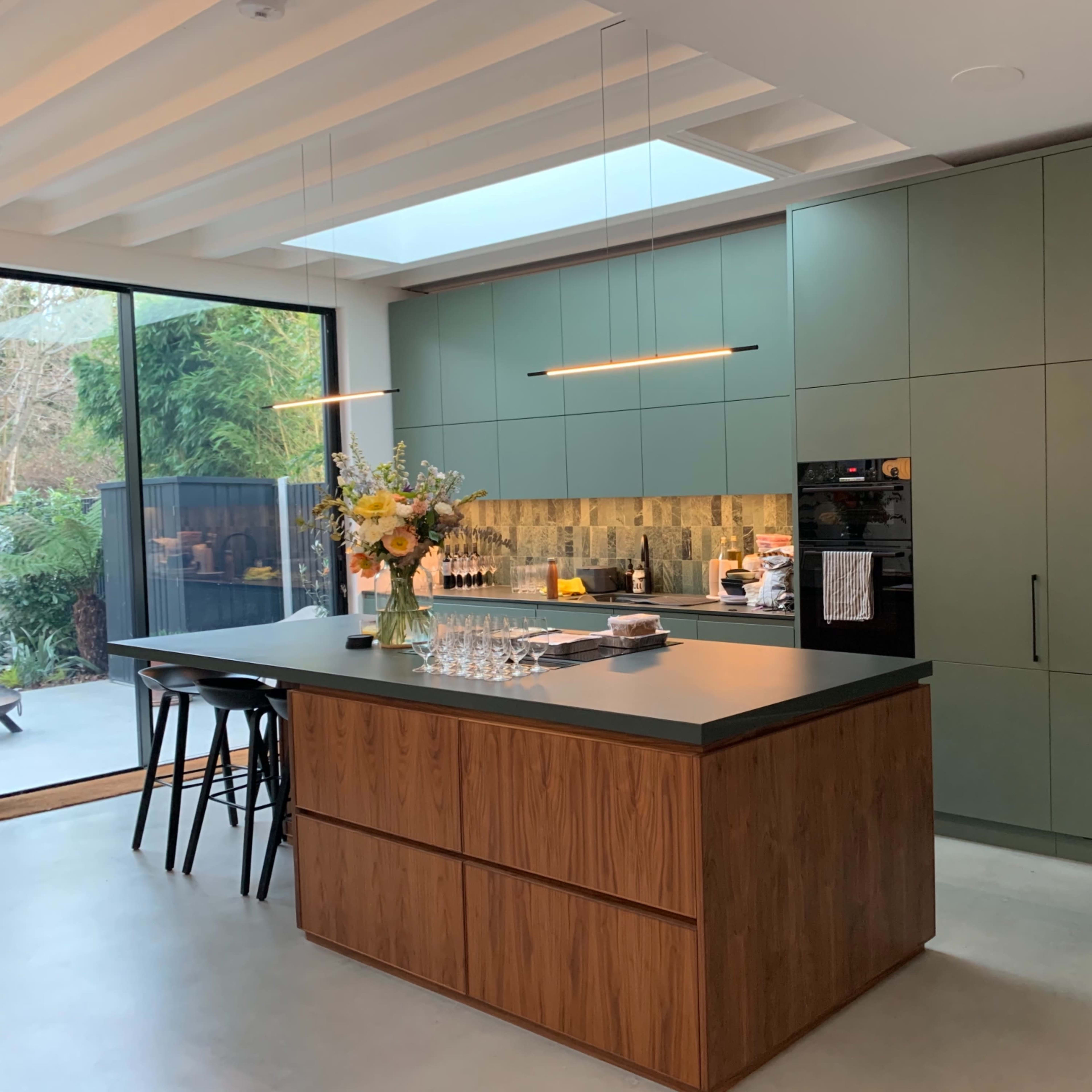 A modern kitchen featuring green cabinets, a wooden island, and a view of an outdoor garden through large glass windows.