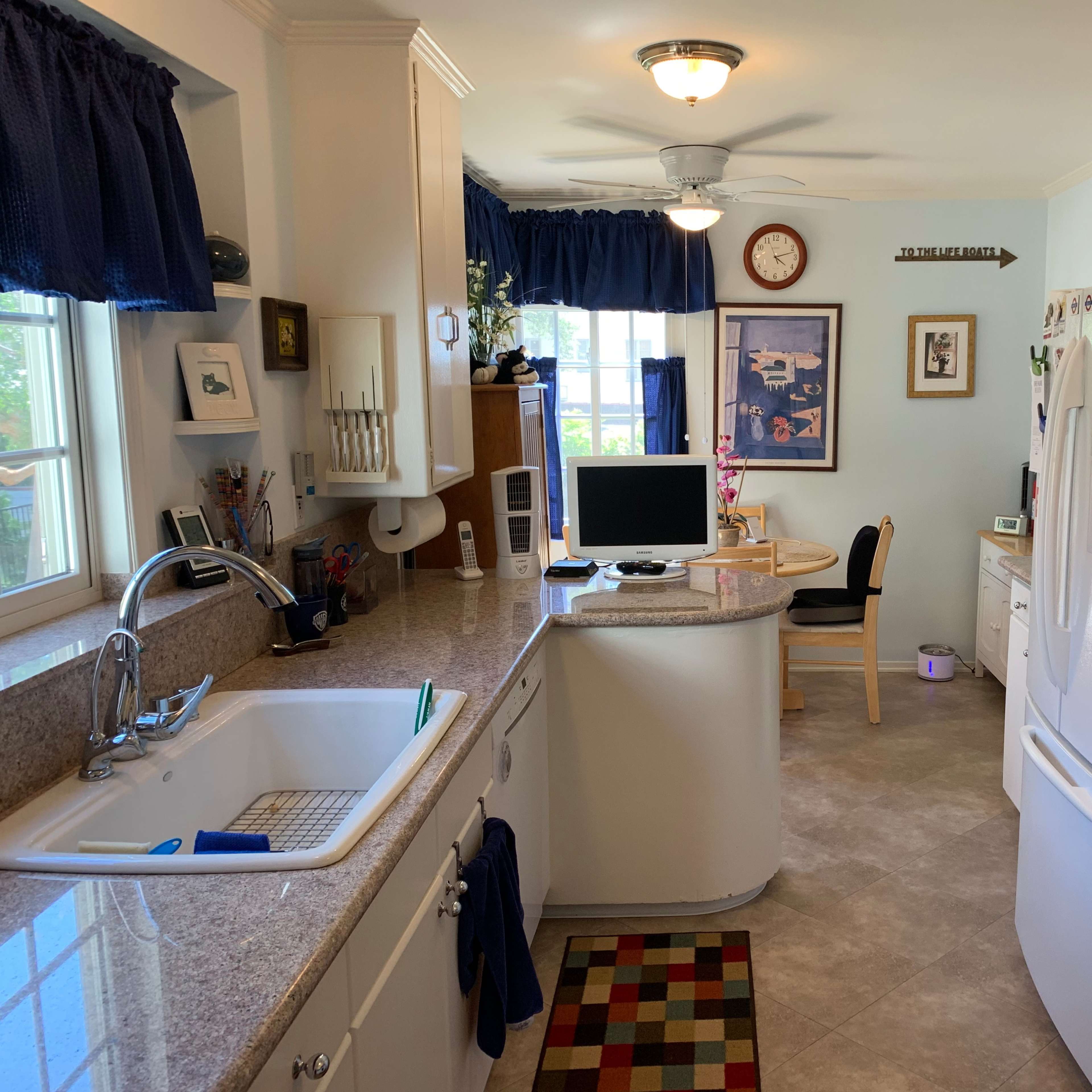 A well-lit kitchen featuring a sink, refrigerator, and a small table with a chair, along with various decorations on the walls and countertops.