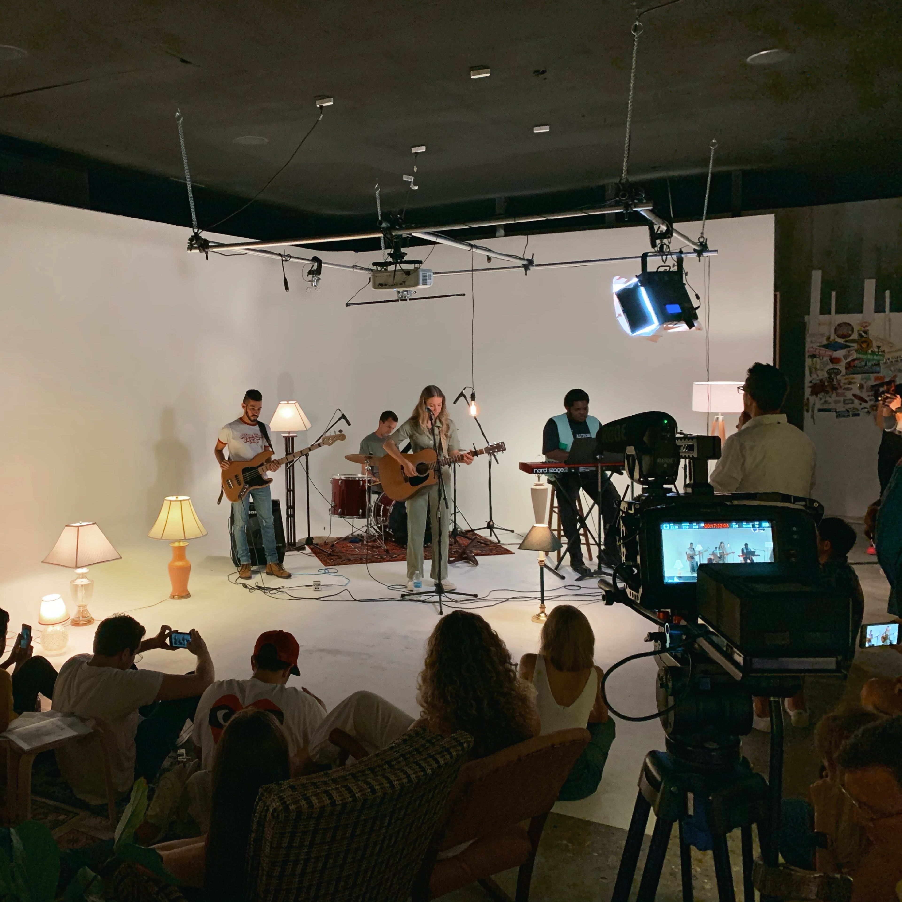A band performs on a minimalist stage with instruments and microphones, while an audience sits on the floor and captures the moment with their phones.
