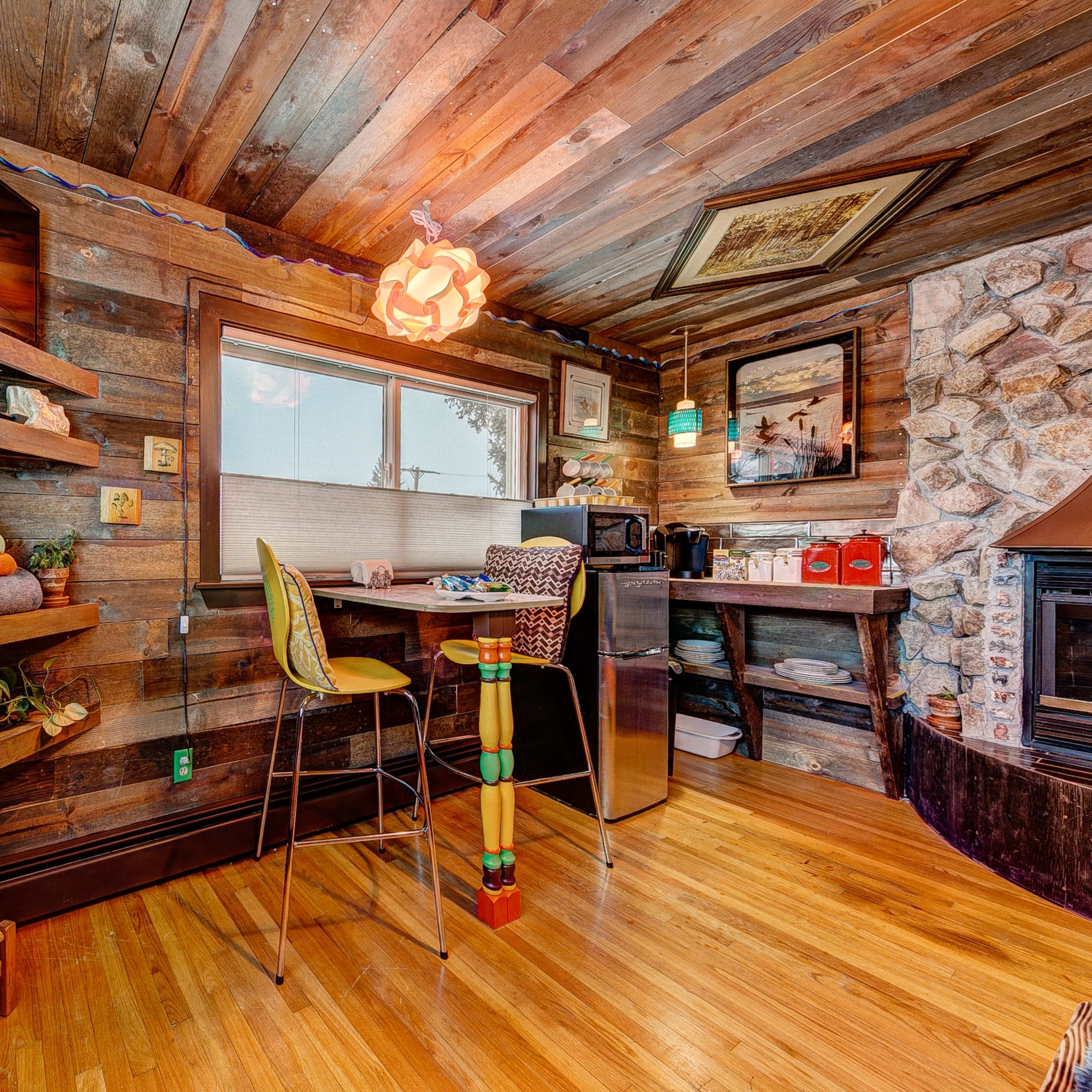A cozy wooden room featuring a small kitchenette with shelves, a microwave, and a bar stool next to a stone fireplace.