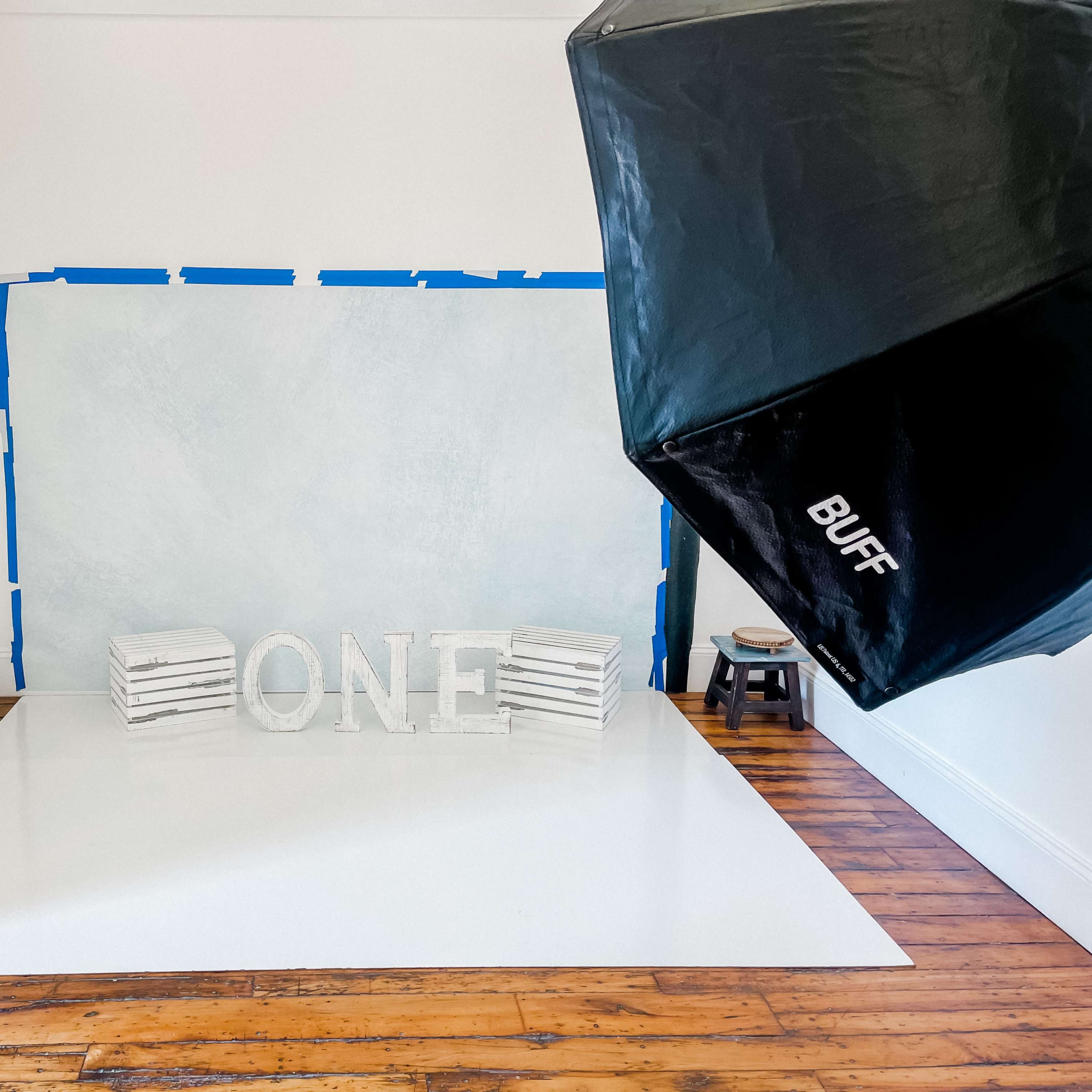 A spacious, well-lit photography studio features a blue-taped backdrop, white floor, and a large, decorative "ONE" displayed in the center.