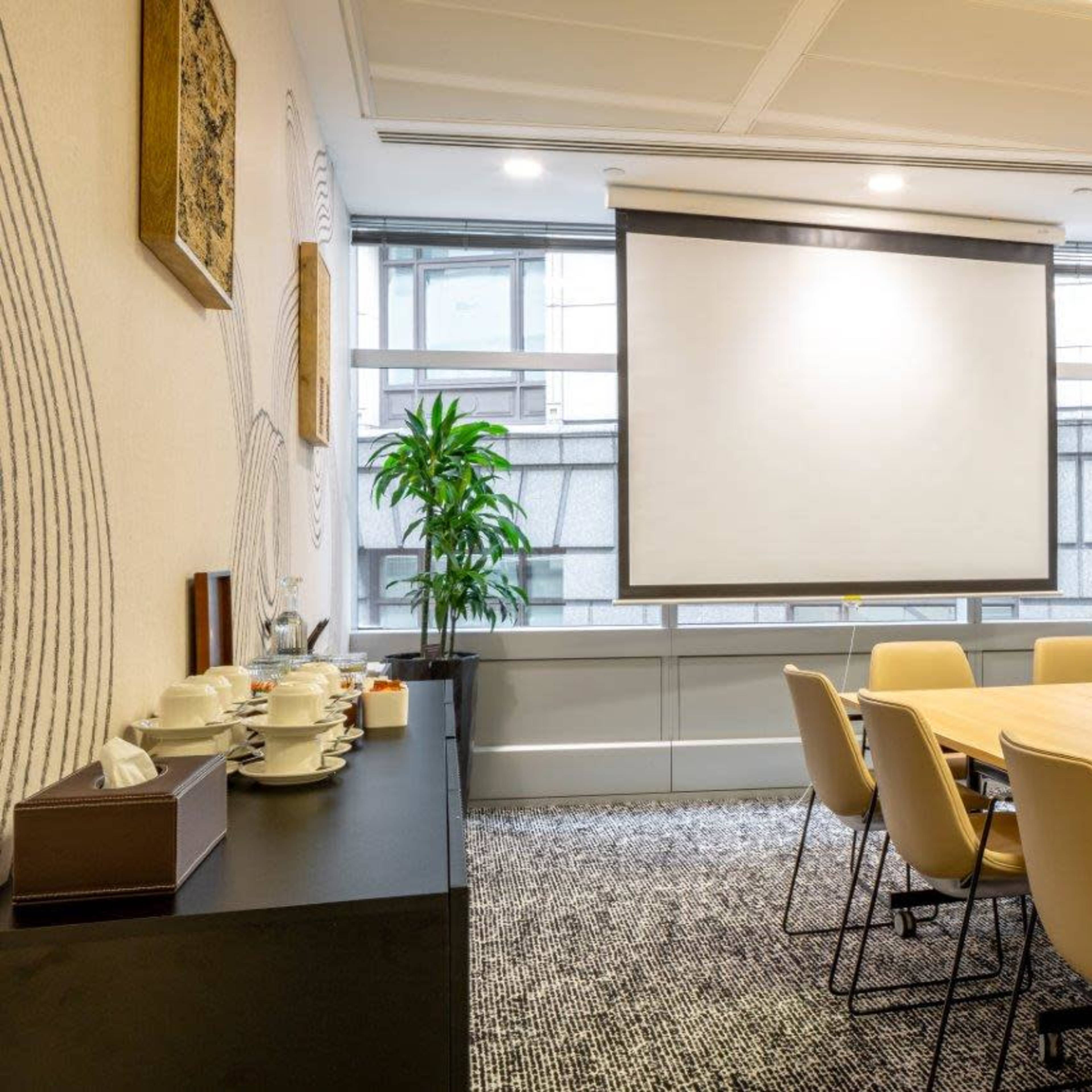 A conference room features a large wooden table surrounded by beige chairs, with a projector screen on one wall and a plant in the corner.