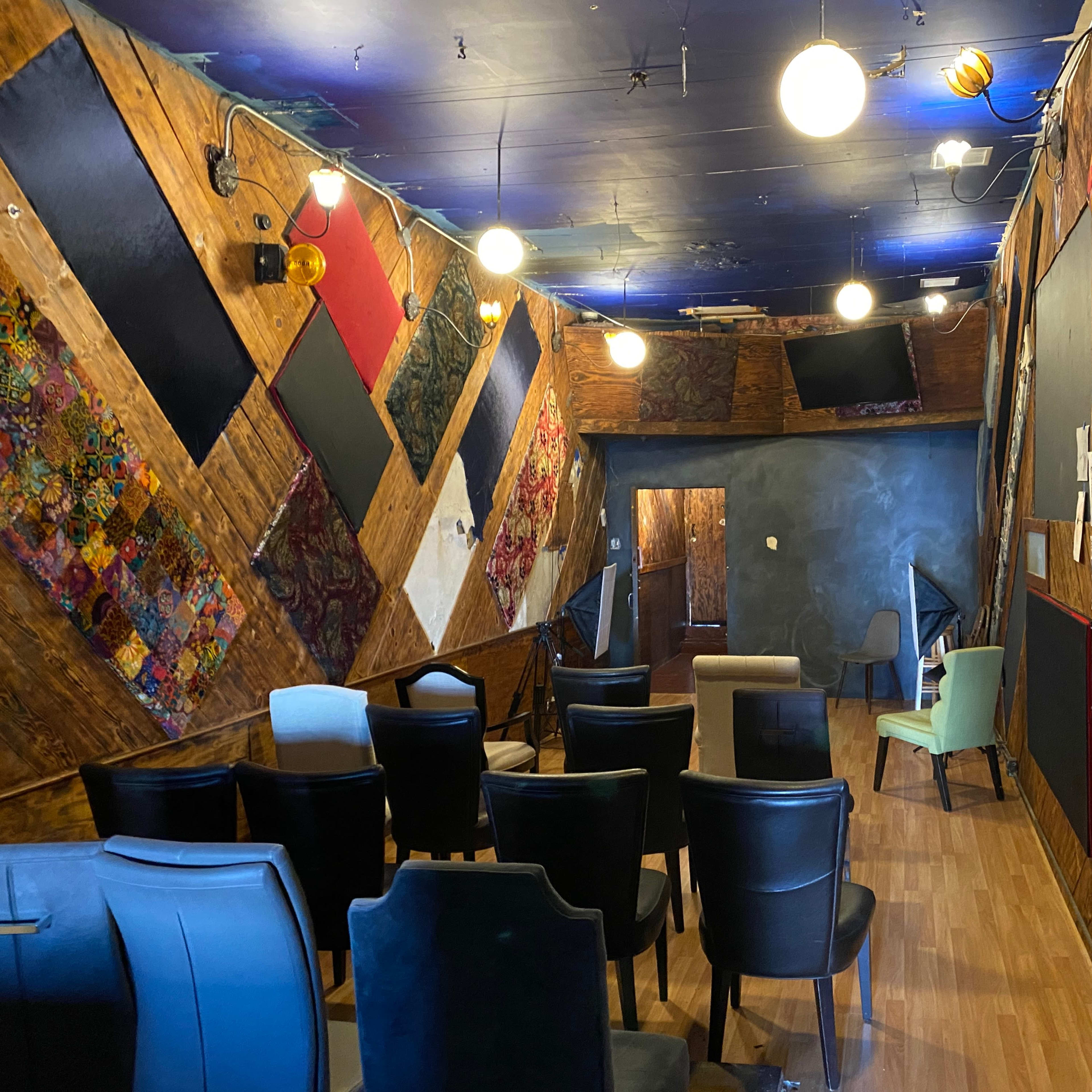 A sparsely furnished room with wooden walls adorned with colorful fabric panels, several chairs arranged in rows, and various lights hanging from the ceiling.
