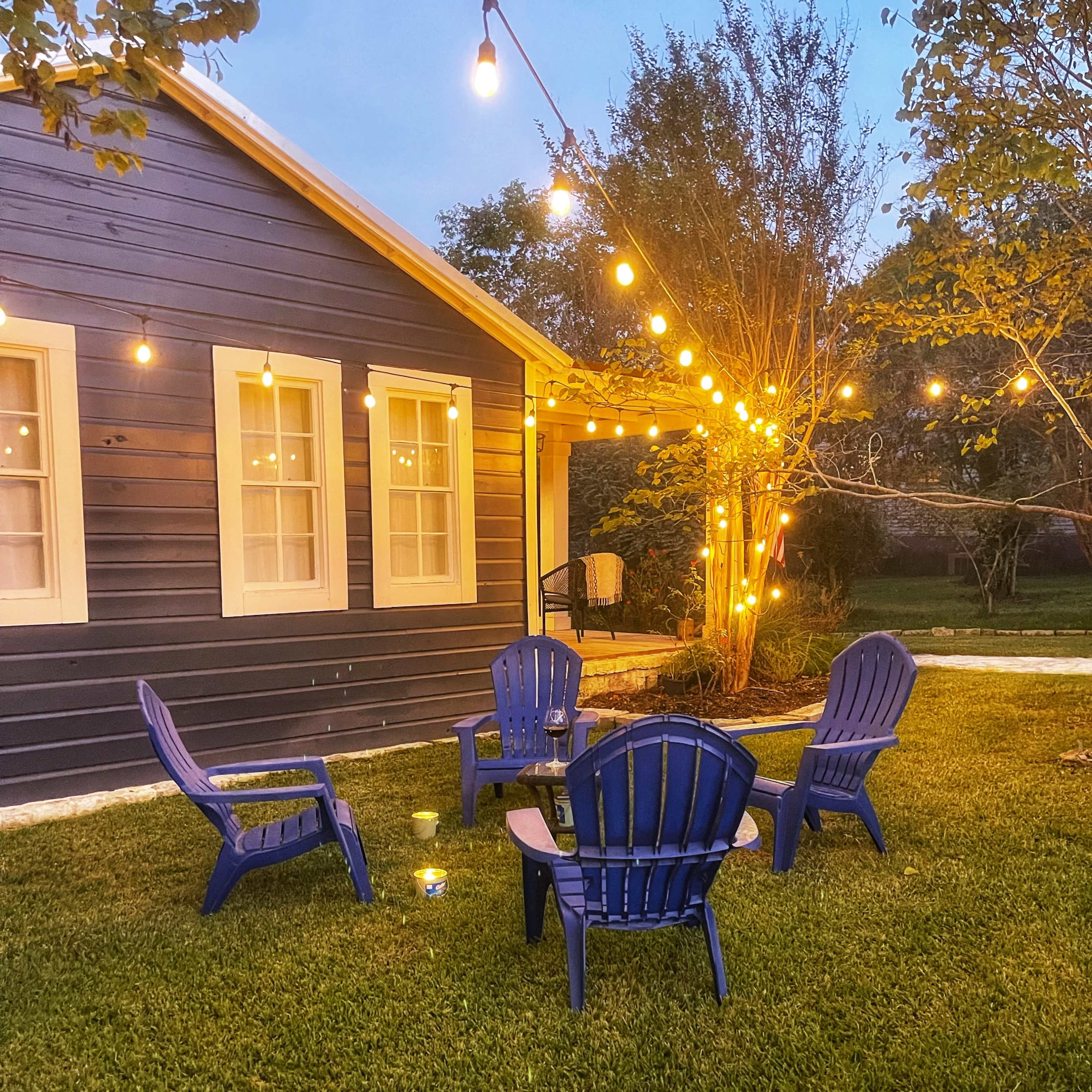 A cozy outdoor seating area features four blue Adirondack chairs arranged in a circle under string lights near a blue house.