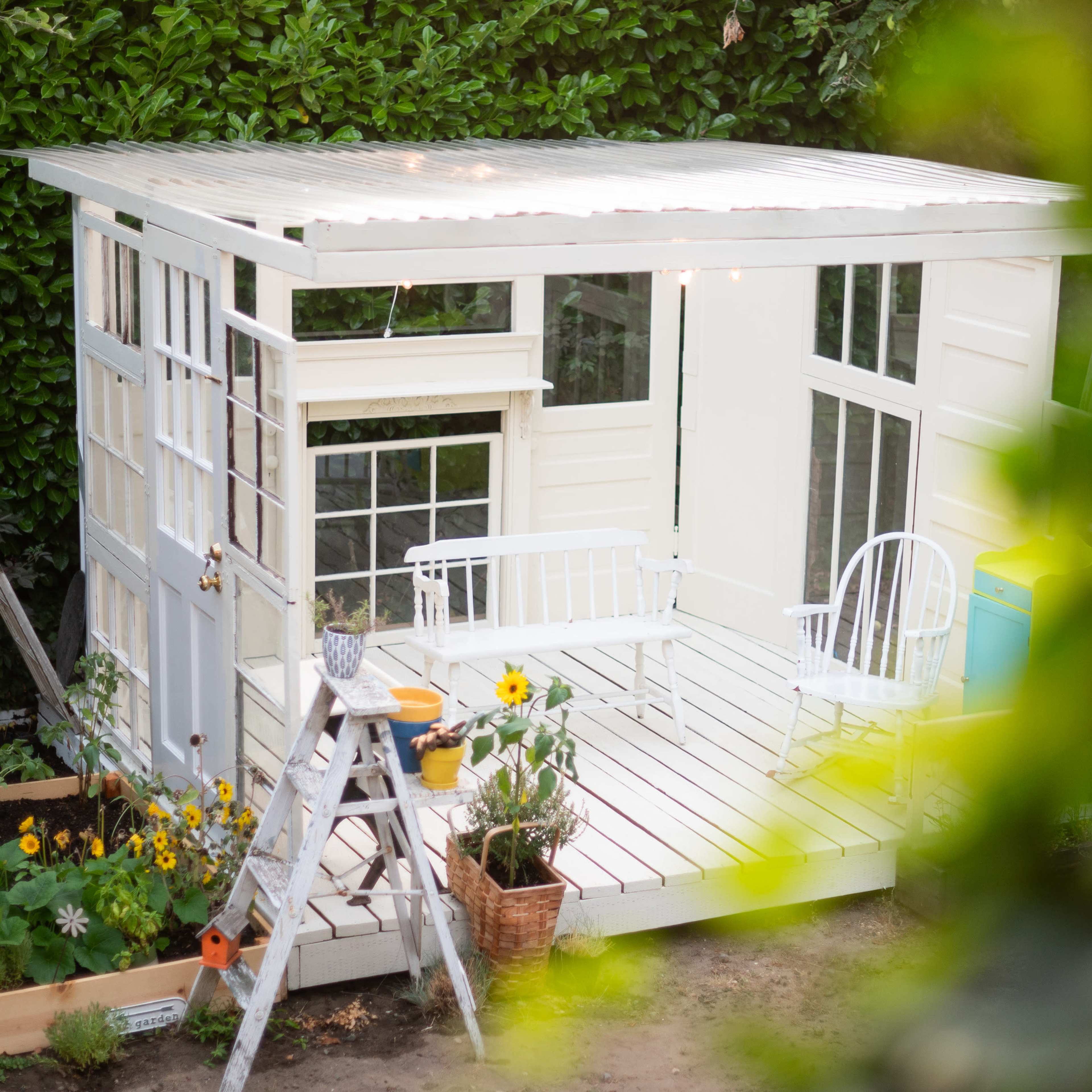 A small white shed with large windows is surrounded by a flower garden and a ladder, with a chair and potted plants on a wooden deck.