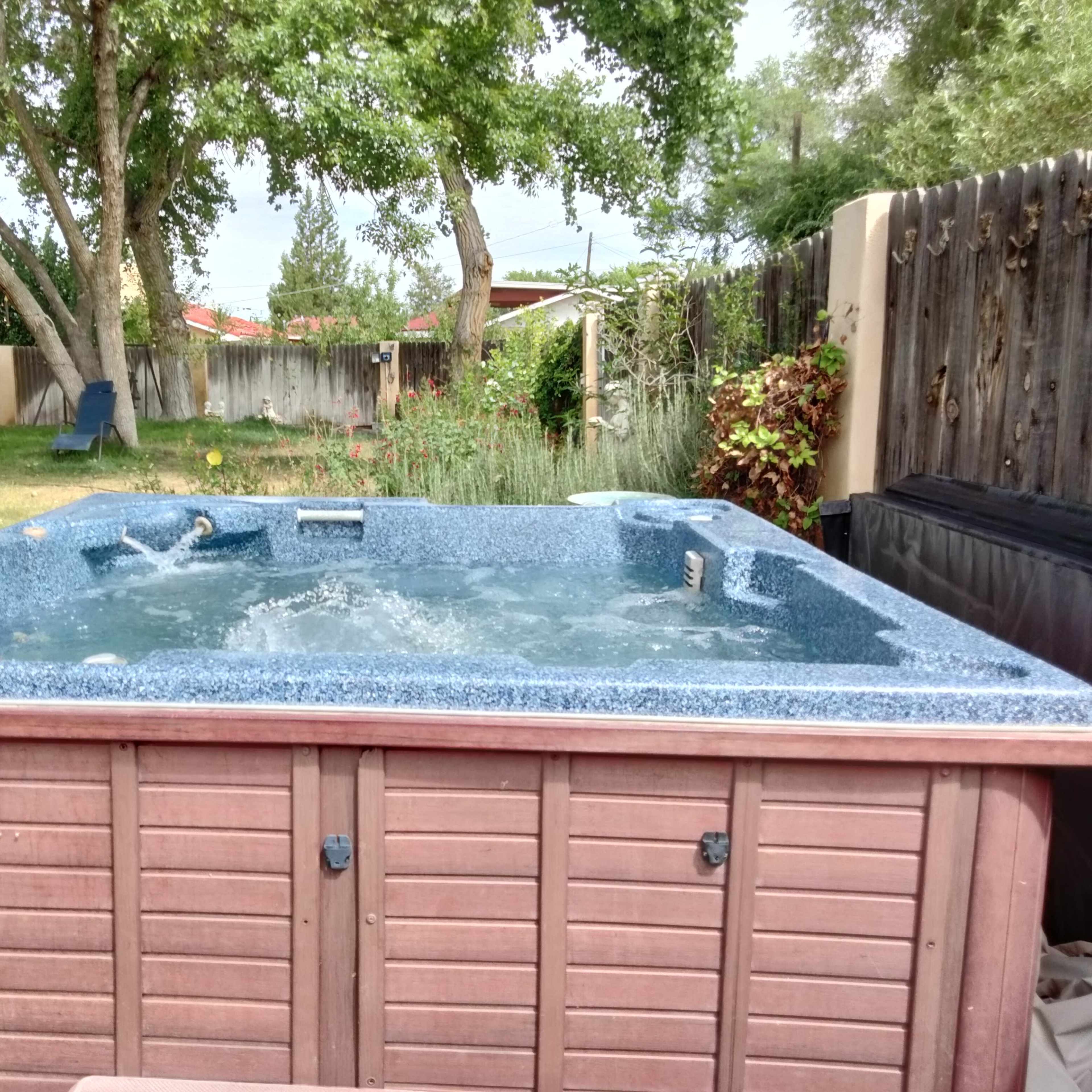 A hot tub with bubbling water is situated in a backyard surrounded by trees and a wooden fence.