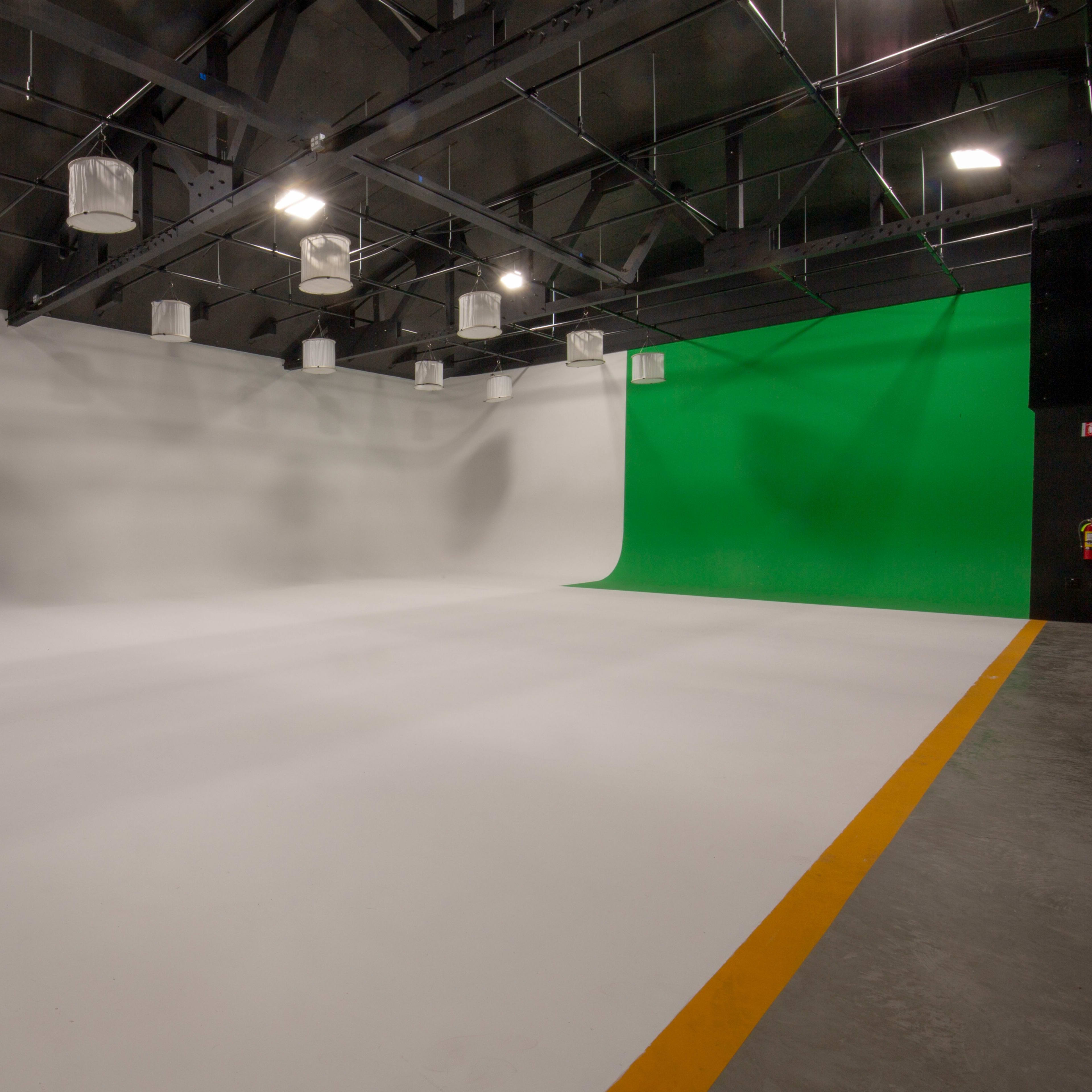 A spacious film studio featuring a white backdrop on one side and a green screen on the other, with overhead lighting fixtures and a yellow border on the floor.