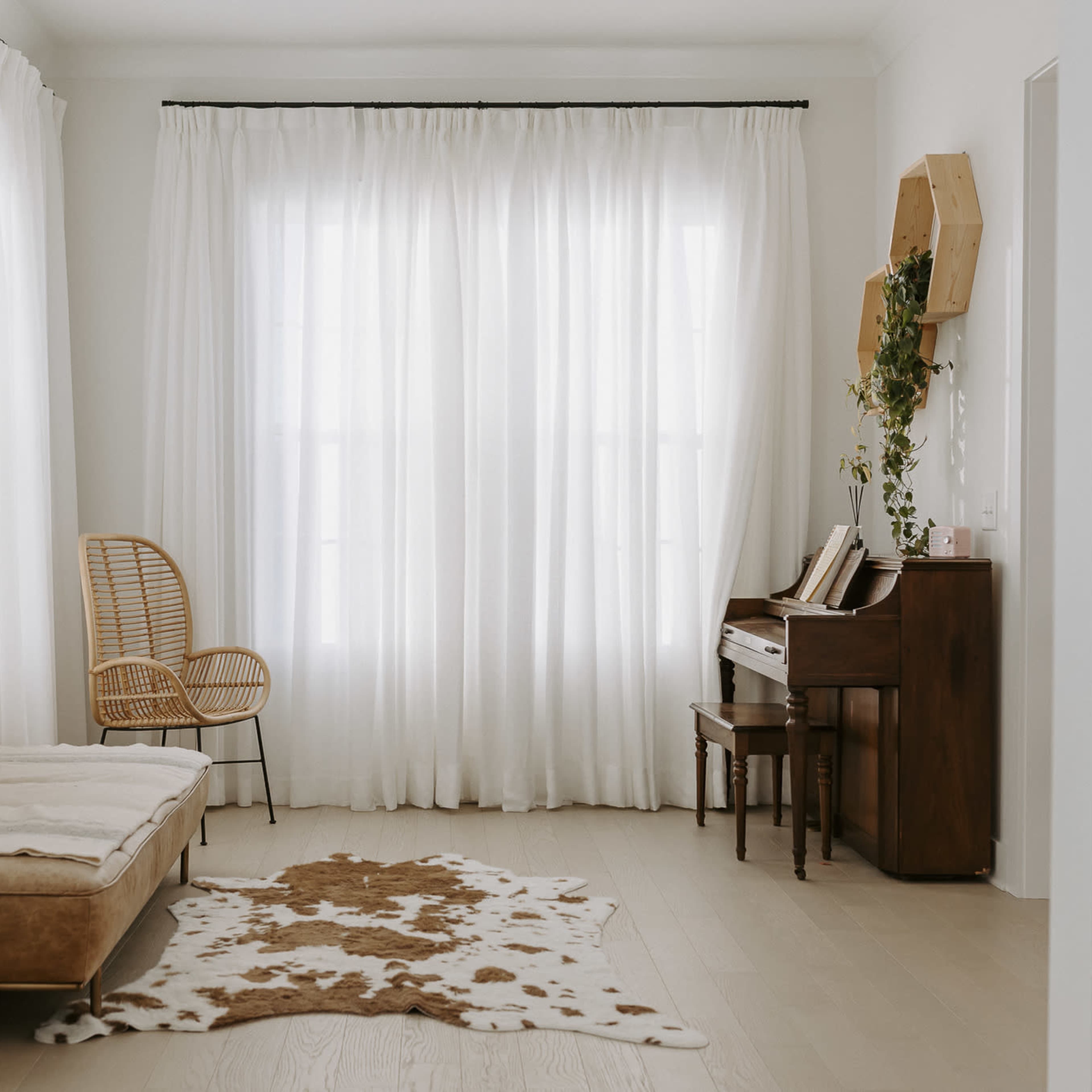 A bright room features large windows with sheer curtains, a wooden piano, a woven chair, and a cowhide rug on the floor.