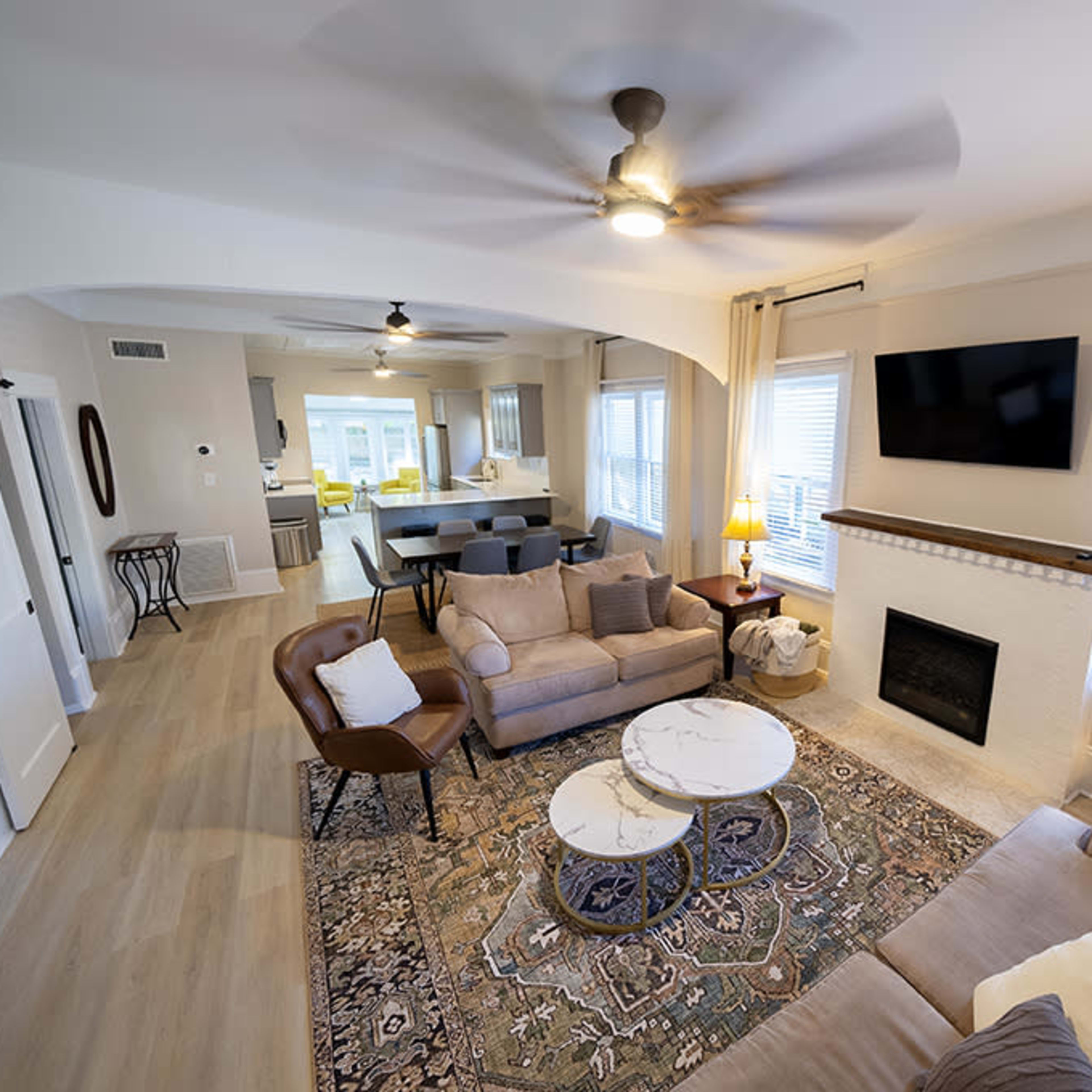 A living room features a beige sofa set, a coffee table on a patterned rug, and a mounted television above a fireplace, with large windows letting in natural light.