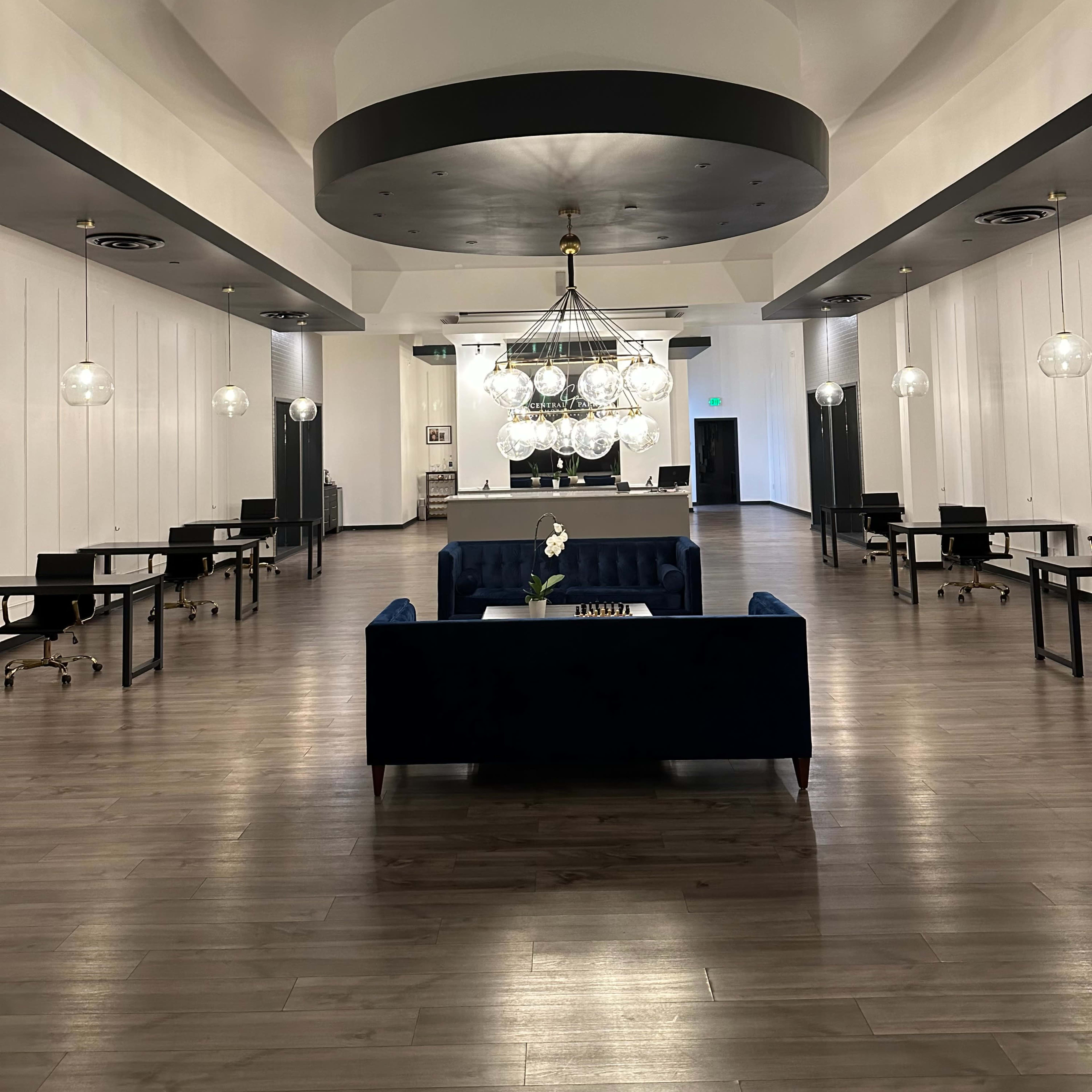 A spacious, modern interior with a central chandelier, seating area, and multiple workstations arranged symmetrically.
