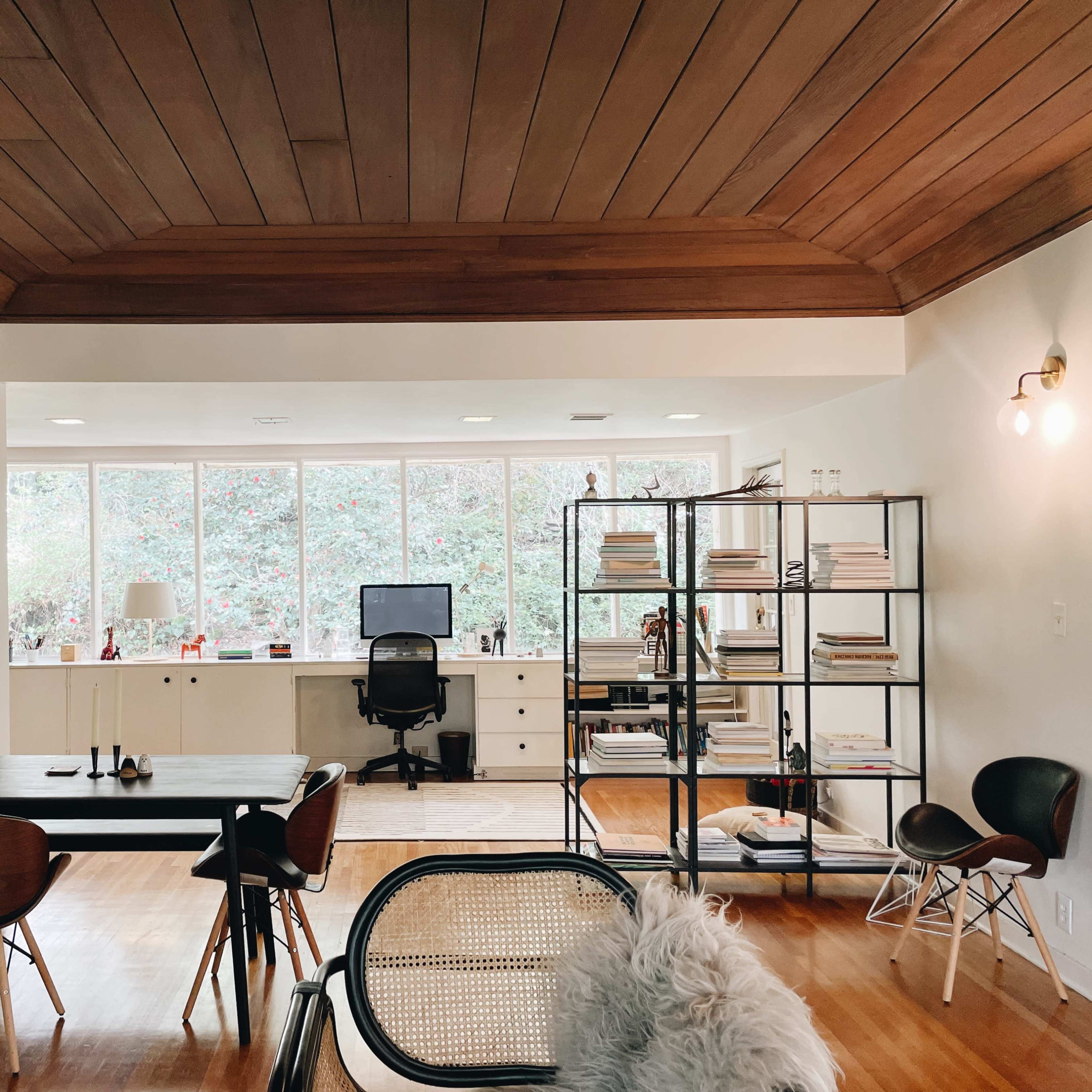 A modern home office with a large window, a desk and chair, and a bookshelf filled with neatly arranged books.