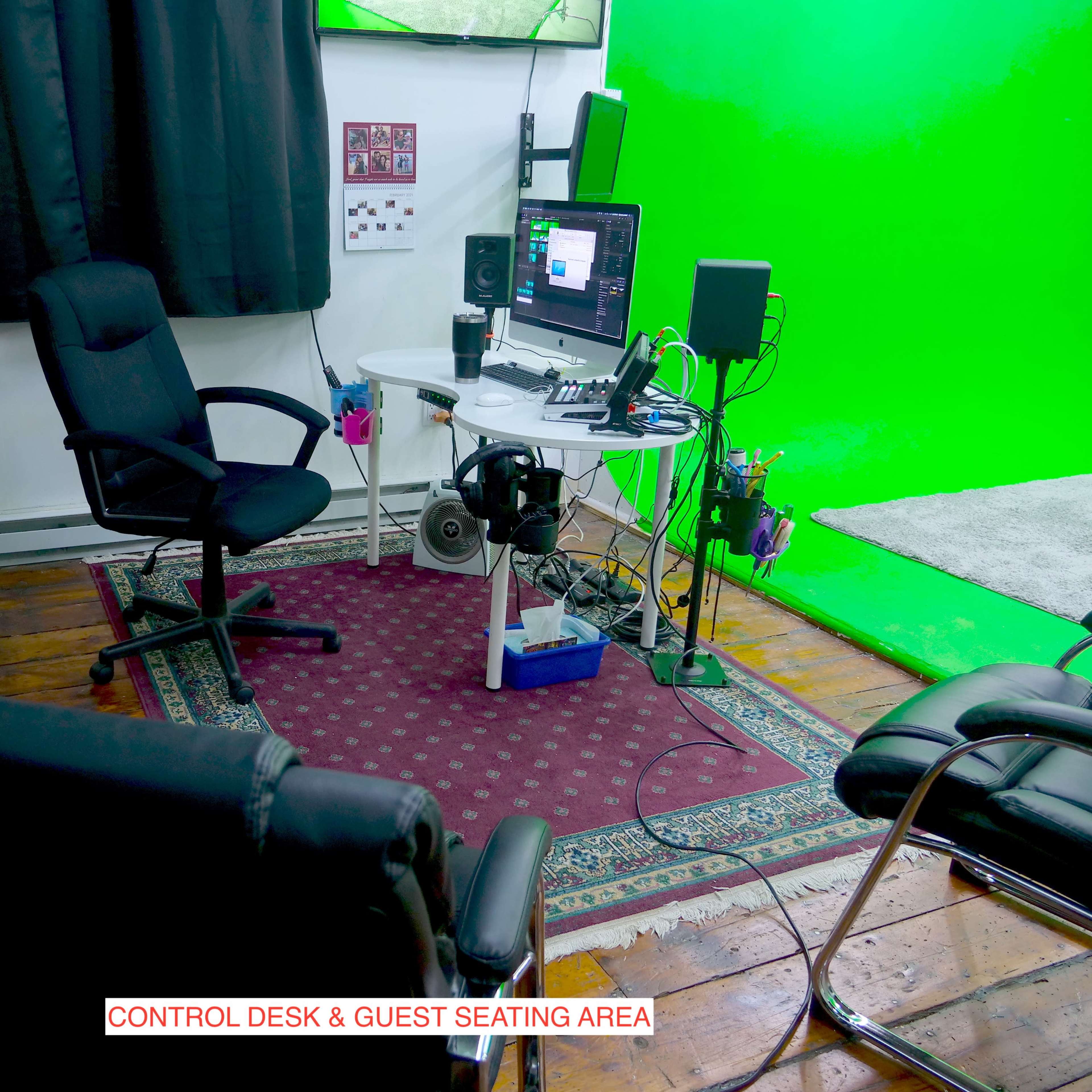 A control desk with computer equipment surrounded by seating in a room with a green screen backdrop.