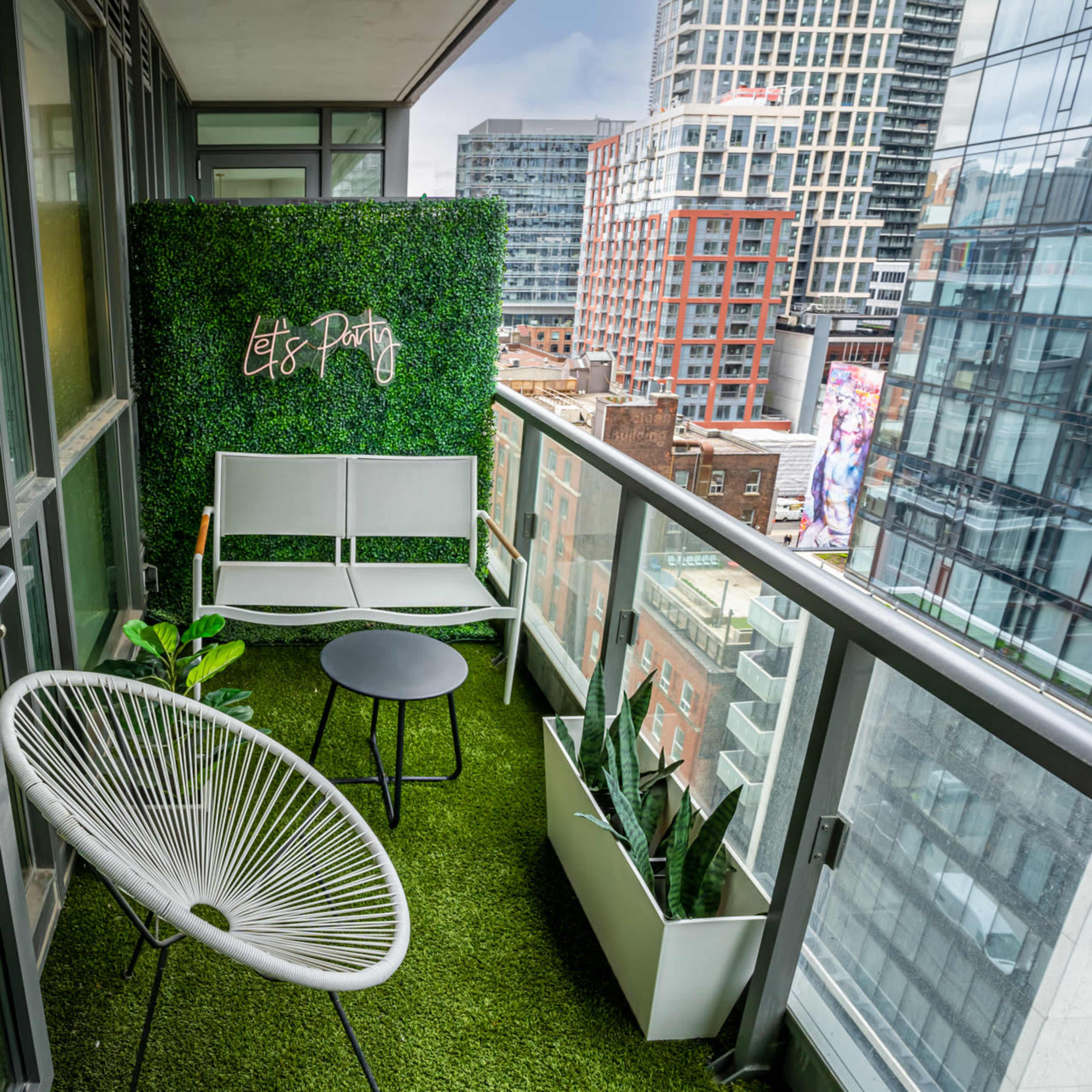 A modern balcony features two white chairs, a small black table, artificial grass flooring, and a green wall adorned with the phrase "Let's Party."