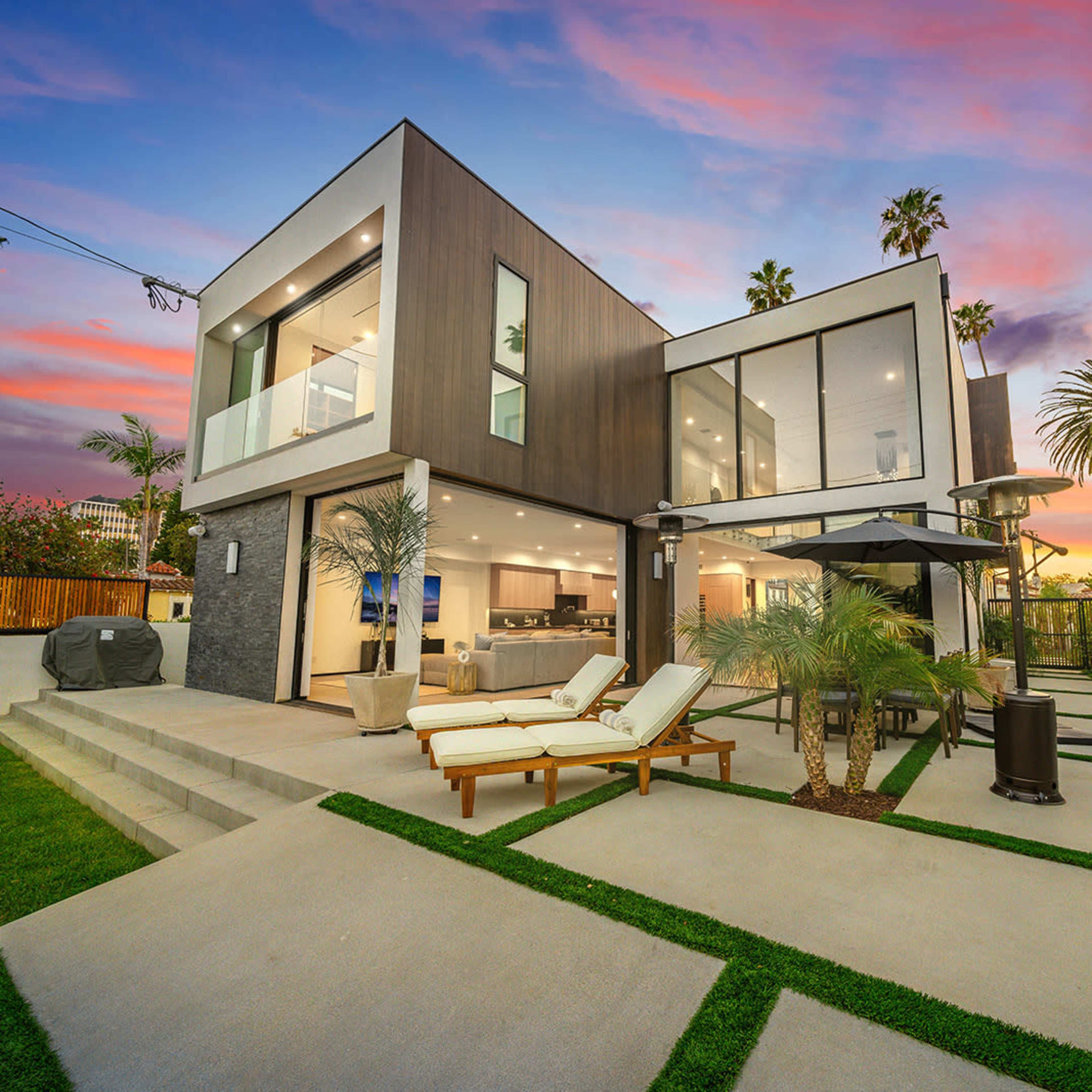A modern two-story house with large glass windows and a landscaped backyard is set against a colorful sunset sky.