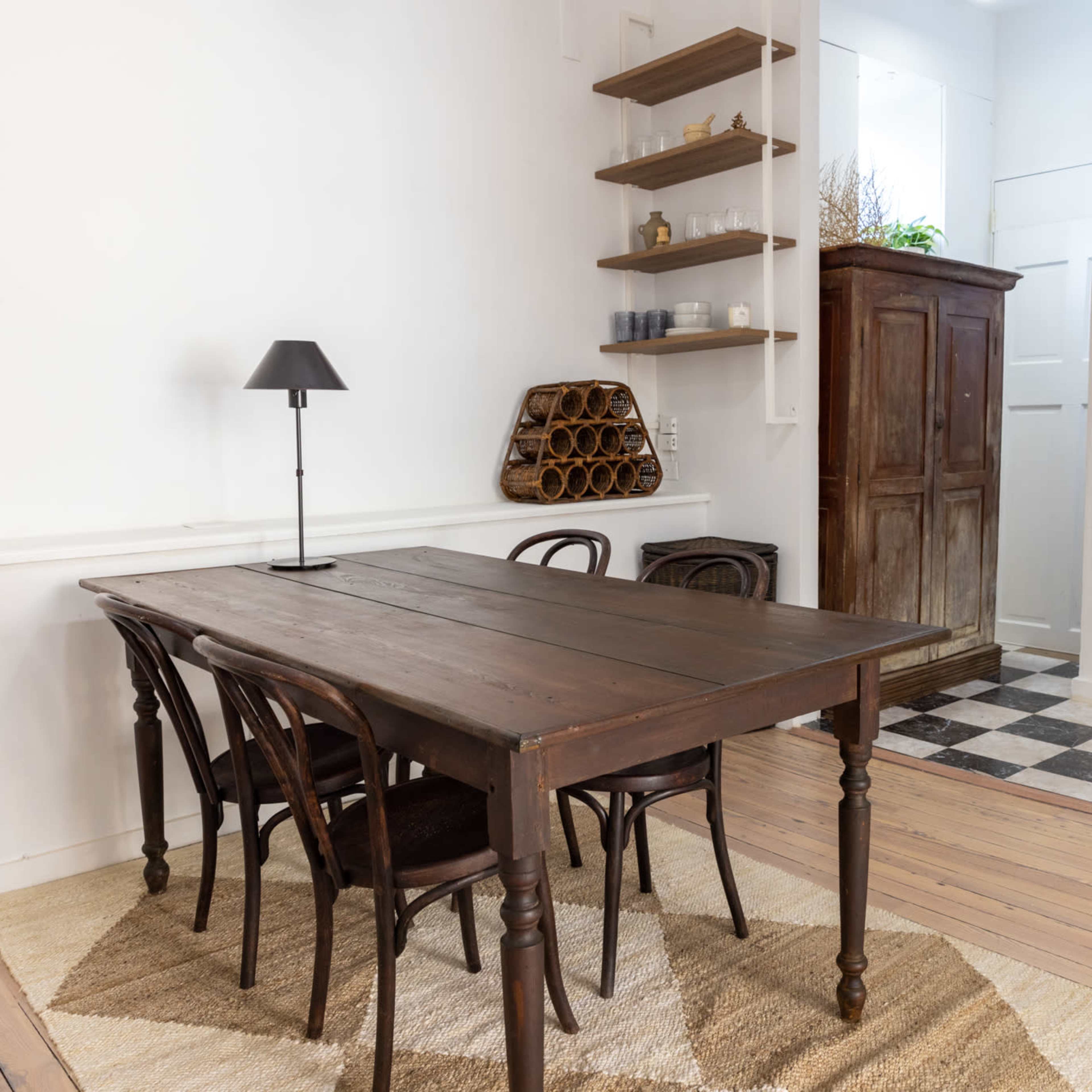 A wooden dining table with four chairs sits on a rug in a room with white walls, featuring a small lamp and shelves on one side, and a patterned floor on the other.