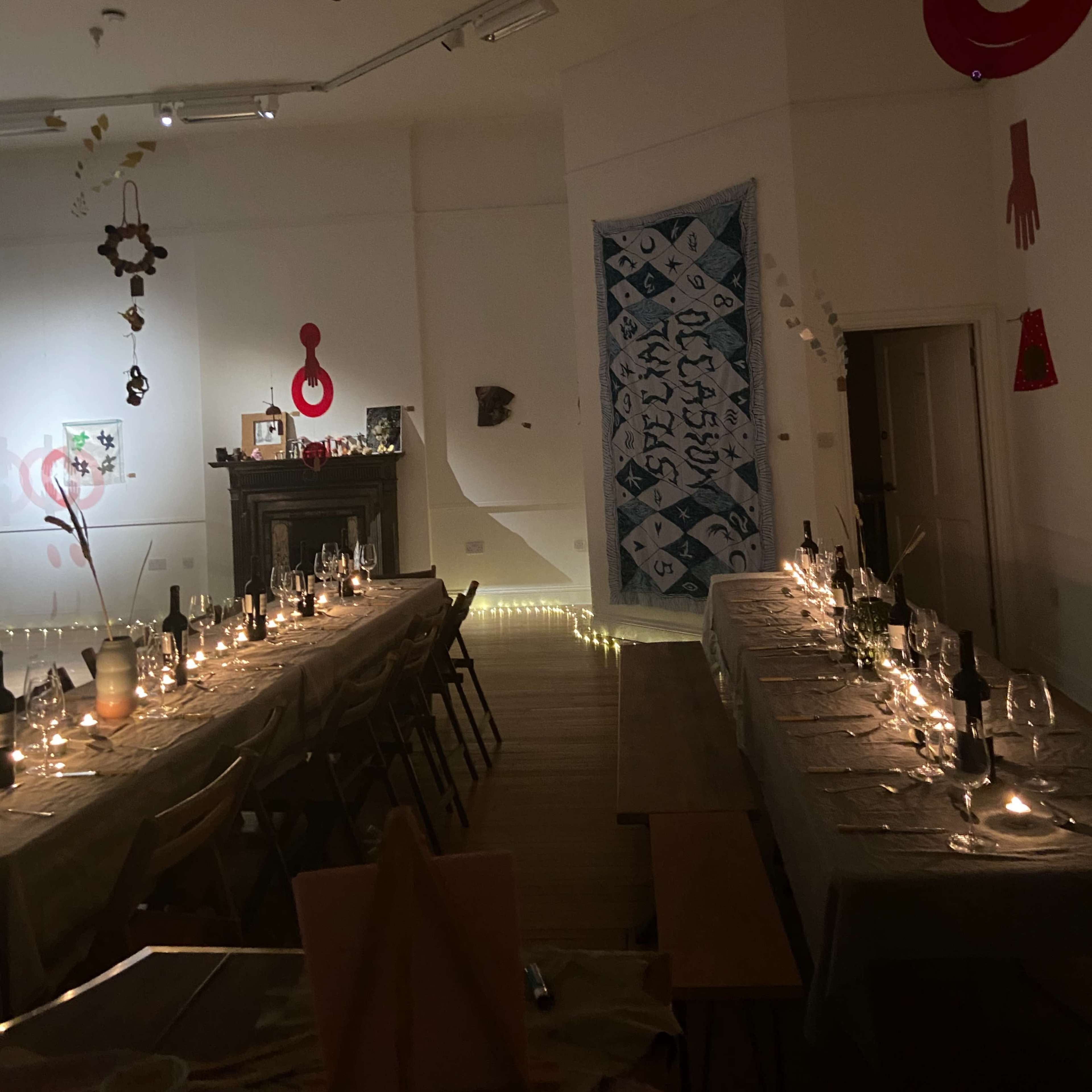 A long dining table is set with candles and wine bottles under dim lighting, surrounded by decorative art pieces on the walls.