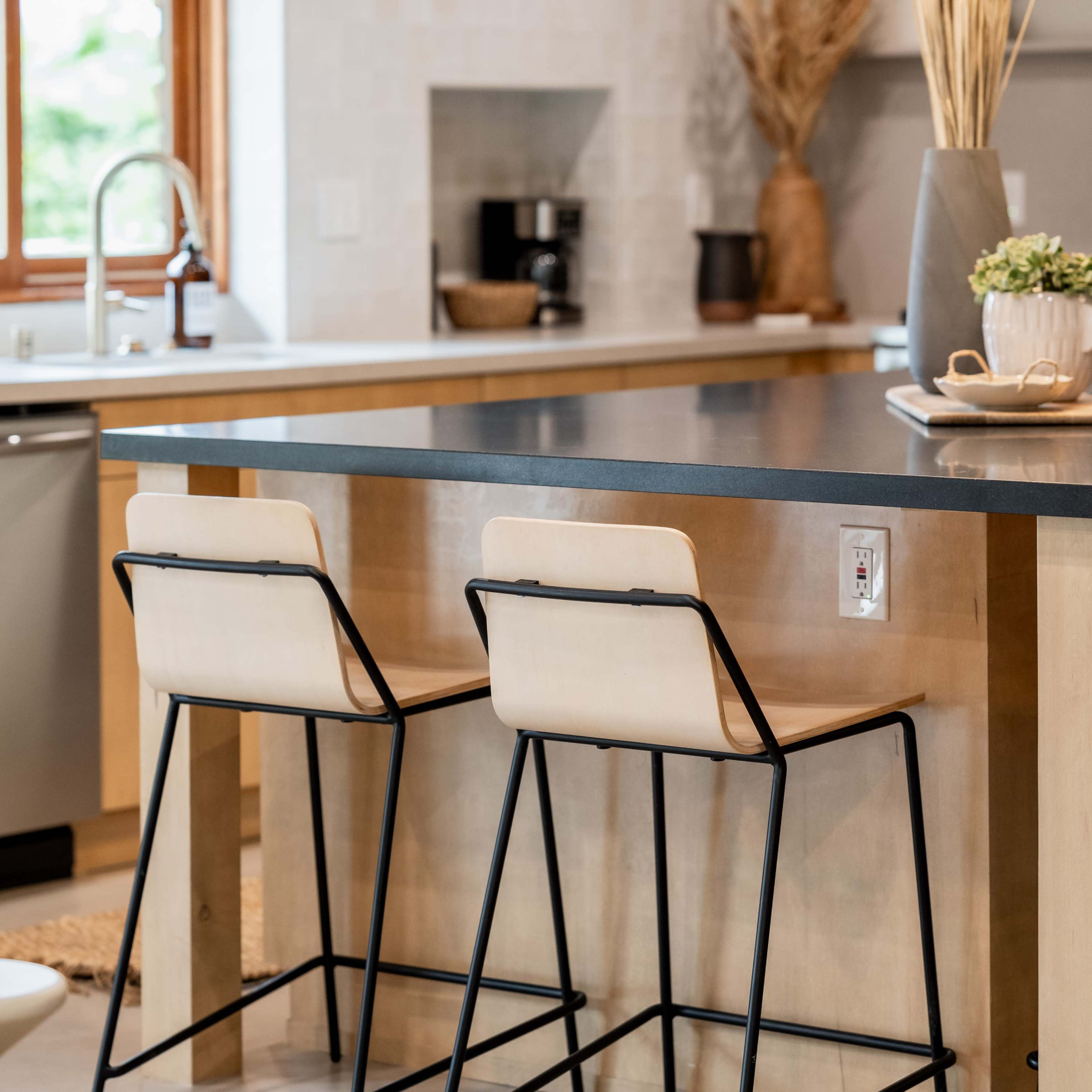 A modern kitchen with a wooden island featuring two black metal and beige cushioned bar stools, a sink, and a coffee maker in the background.